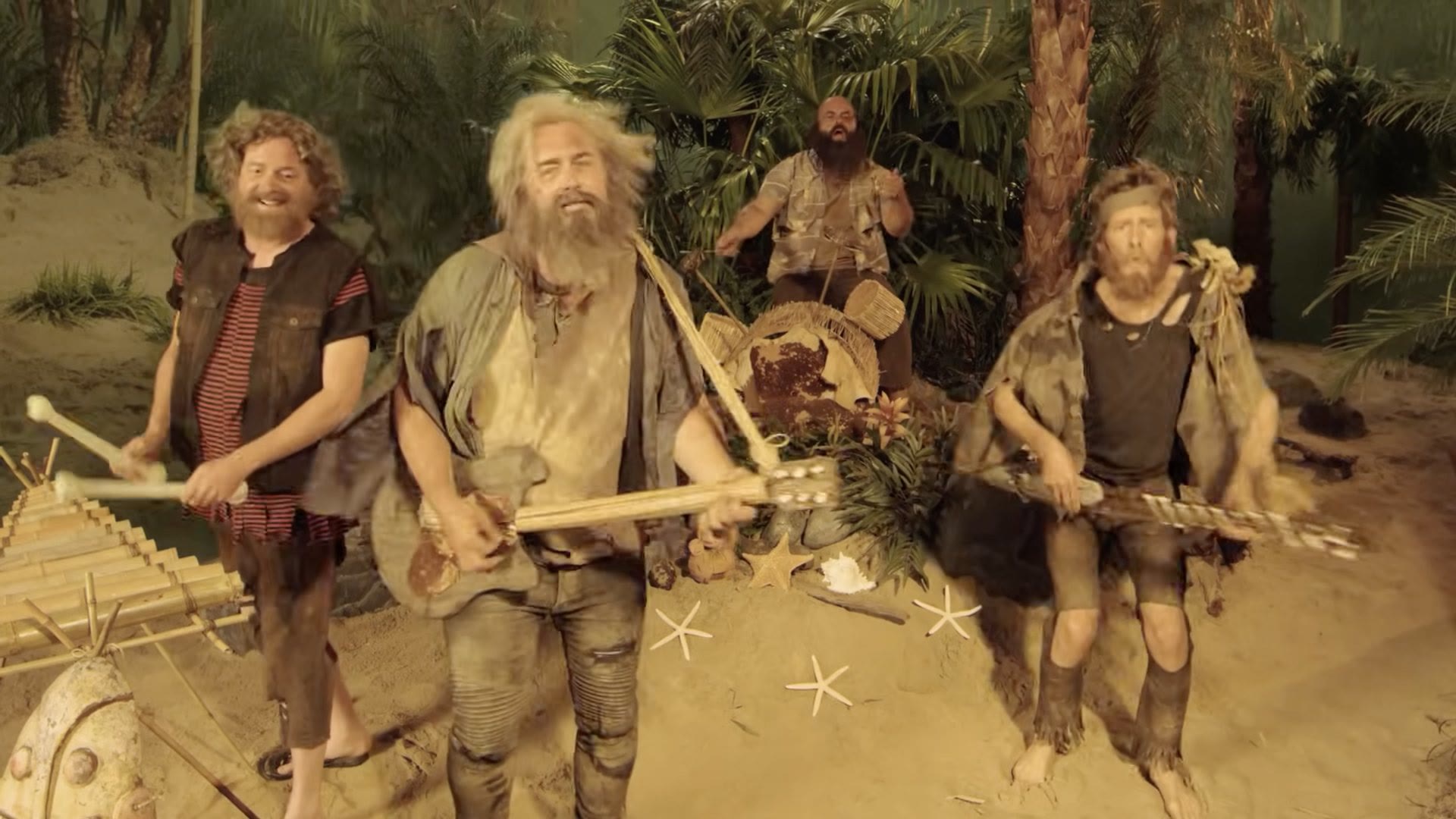 A frame from the finished video: the four band members as castaways on the island set, playing instruments improvised from bamboo and driftwood, with starfish and shells dressed into the sand.