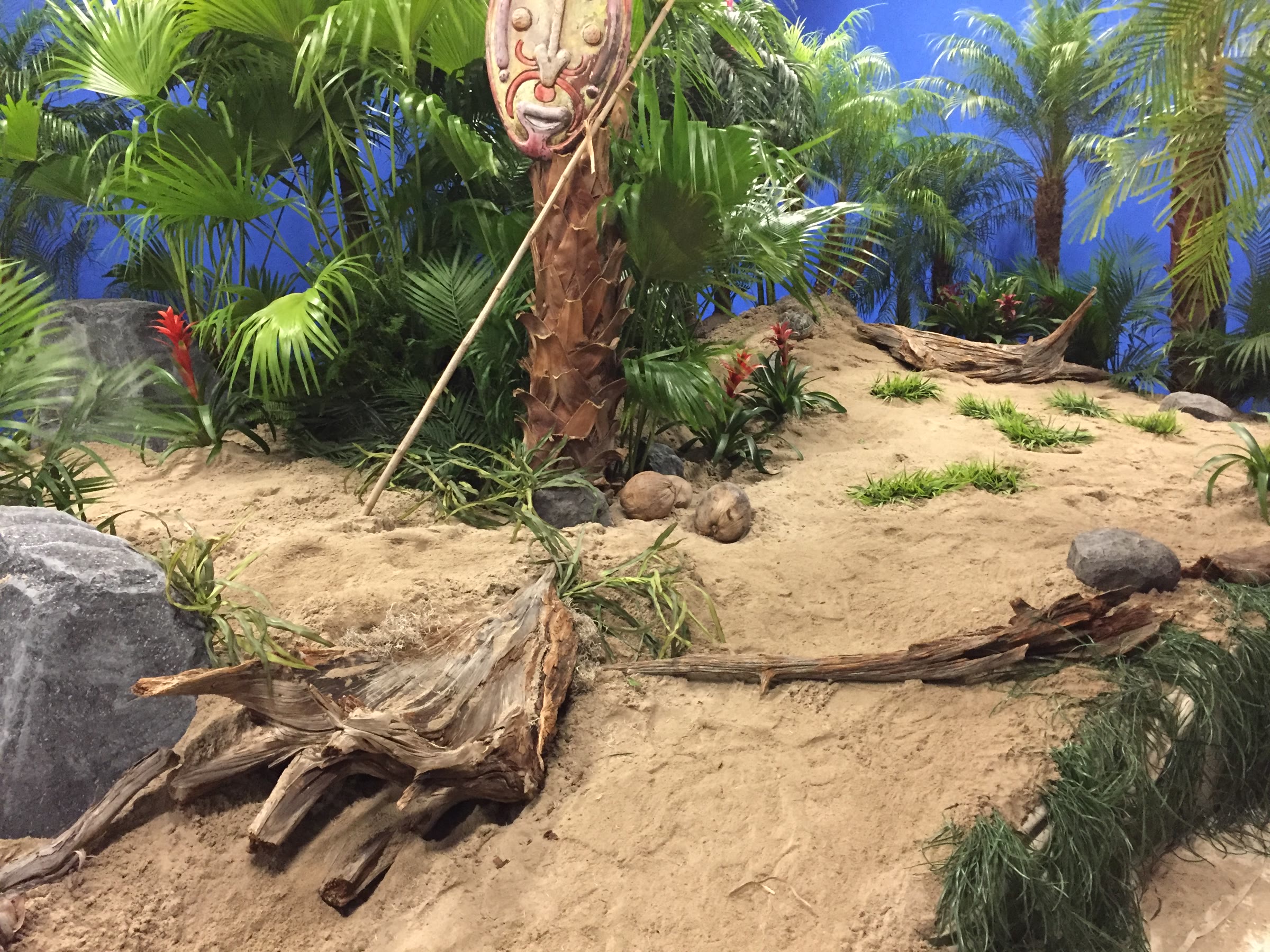 The finished island set: a sand dune planted with palms and red bromeliads, weathered driftwood and grey rock forms across the foreground, a carved mask mounted on a post beside a leaning bamboo pole, all against a lit blue backing.