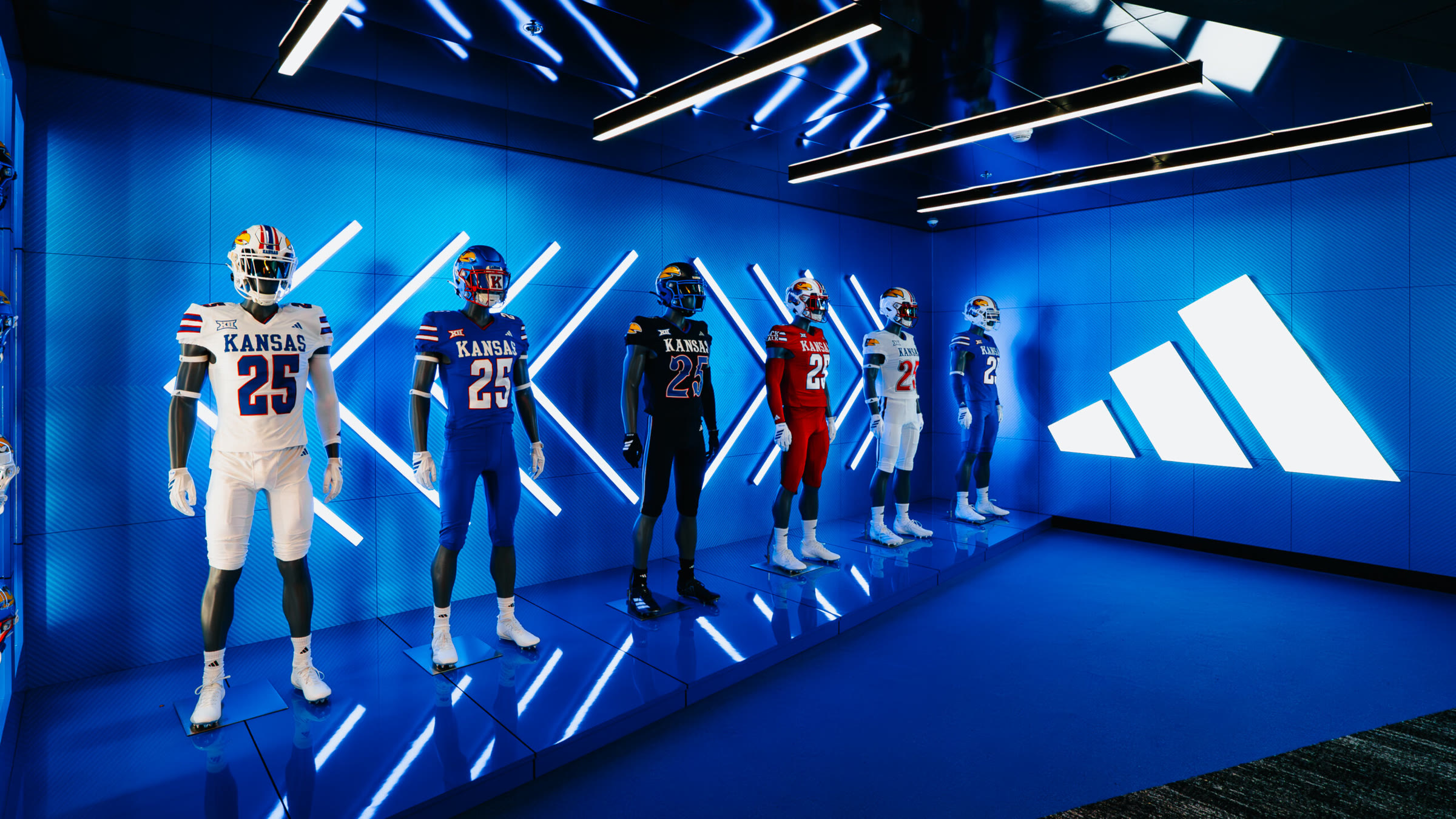 Kansas Football gear display: backlit uniforms in a deep blue mirrored room with X-pattern light walls.