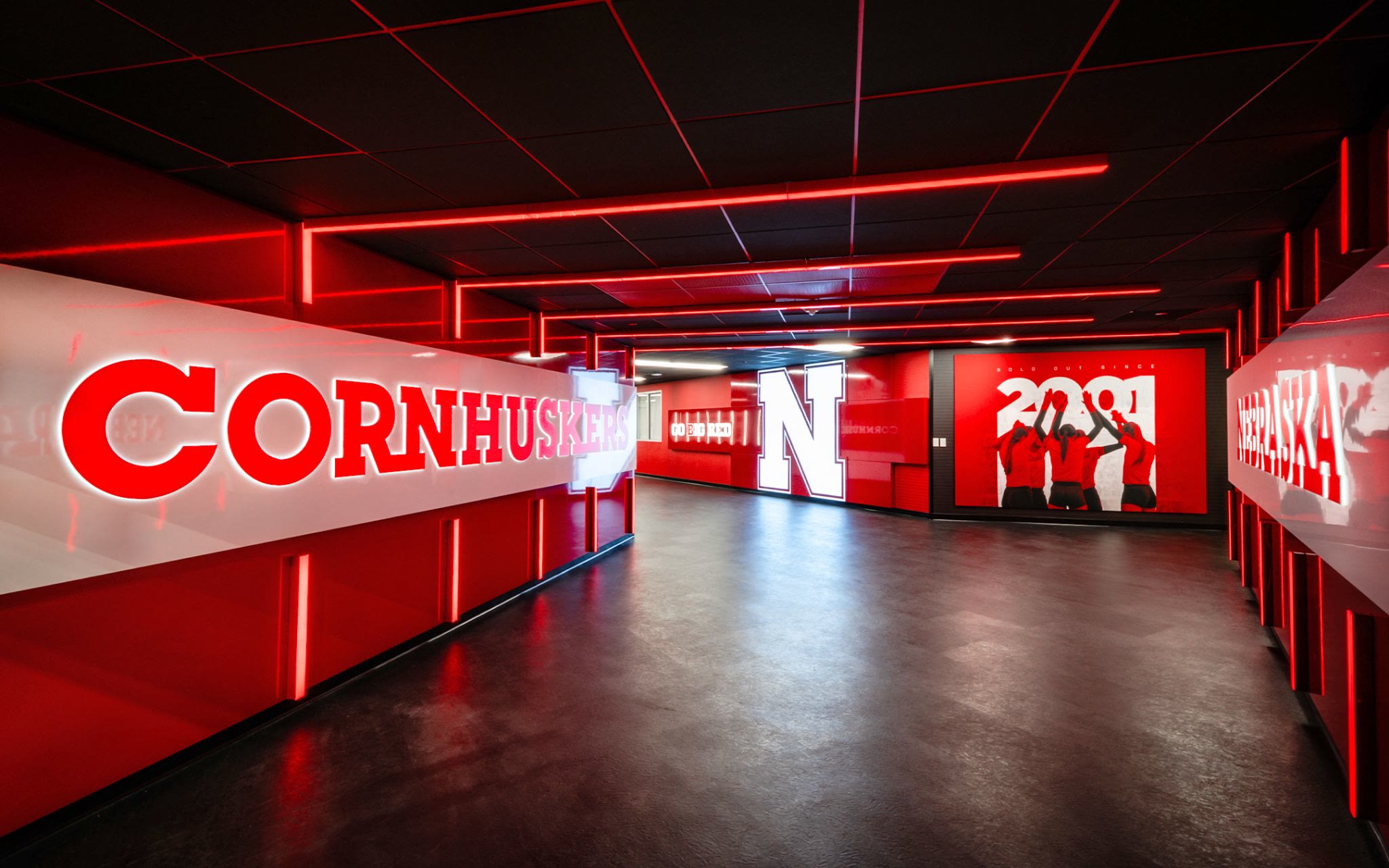 Inside the Nebraska Volleyball player suite: CORNHUSKERS in dimensional red letters down a white corridor wall, red light lines overhead, the lit N mark at the far end.