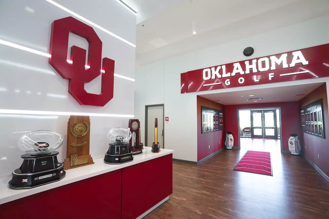 The Oklahoma Golf clubhouse entry: championship trophies on the counter under the OU monogram, the Oklahoma Golf corridor beyond.