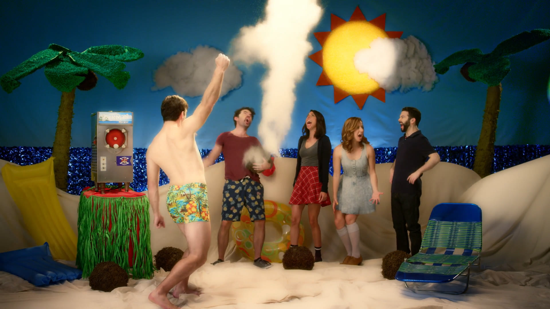The beach cold-open: a deliberately fake seaside built from tinsel ocean, a cardboard sun, cotton clouds, piñata-shop palm trees and fabric dunes, with the cast celebrating around a coconut erupting in a white geyser.