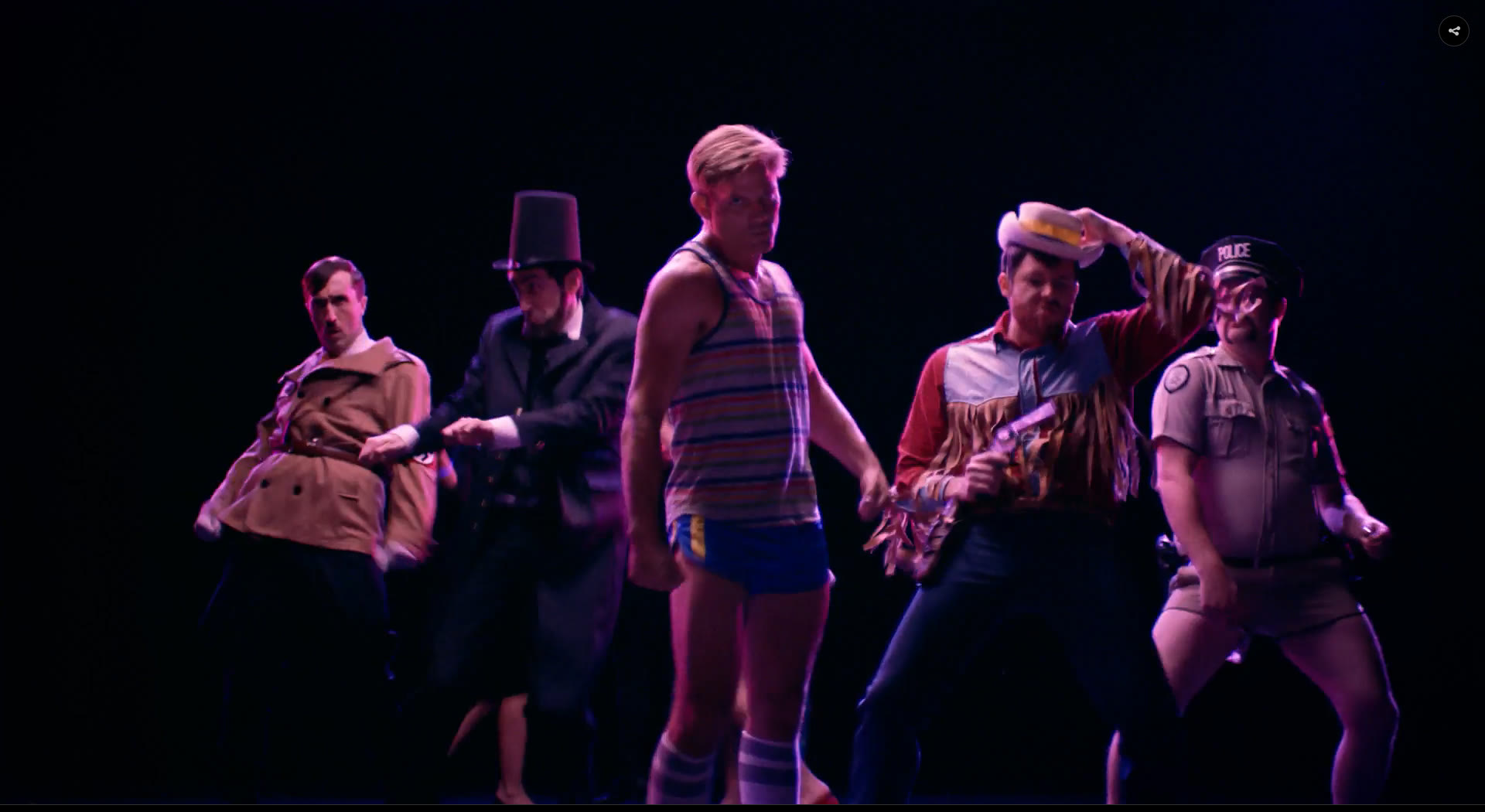 A fantasy dance number on a black void stage: Casper Van Dien front and centre in a tank top, with Lincoln, a police officer and a cowboy behind him.