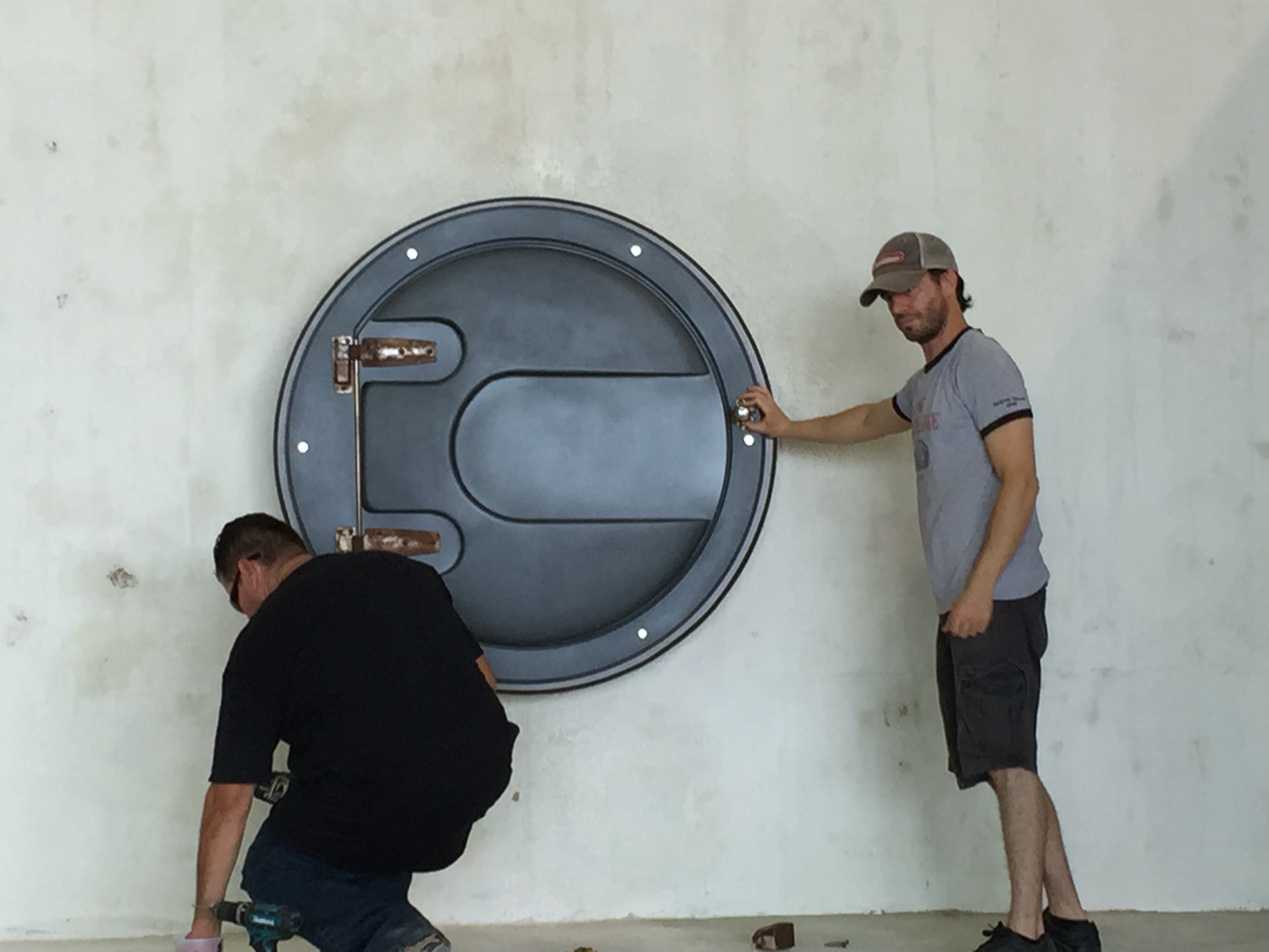 Two crew hanging the flat hatch on a bare wall — one steadying it at the rim, one kneeling with a cordless driver.