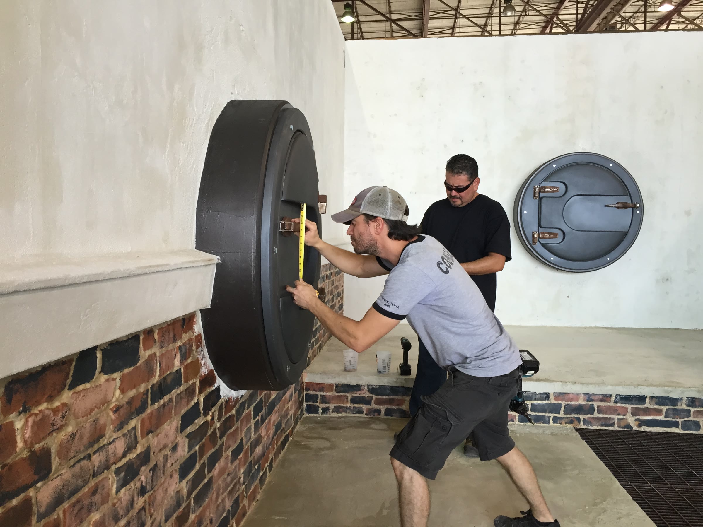 The deep barrel-type hatch being tape-measured into position while a second crew member steadies it; a finished flat hatch is already hung on the wall behind.