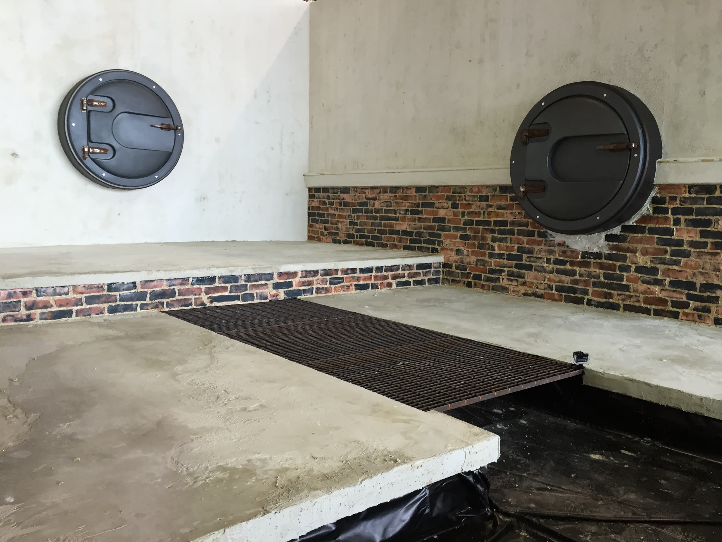 Two hatches installed on adjacent walls above a brick wainscot, with the raised platform and steel floor grate in place; the plaster is still clean and unaged.