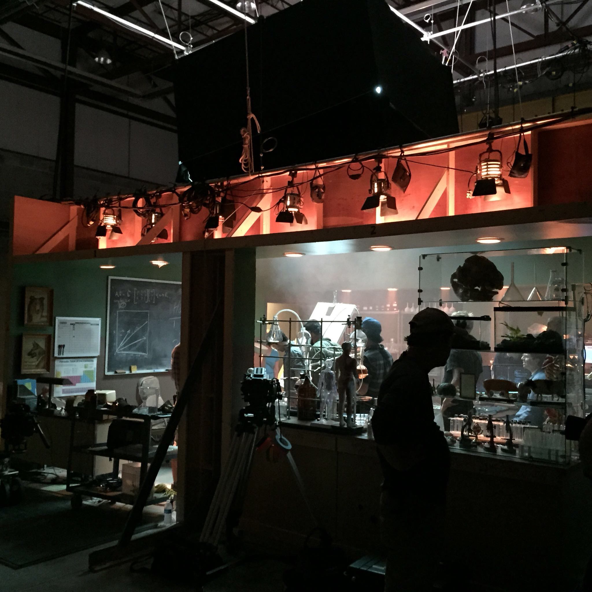 The Lab from the stage floor with the studio around it: practical lights rigged in a row along a raised header above the set, a camera on sticks, a crew member in silhouette, and the dressed glass specimen case lit inside the room.