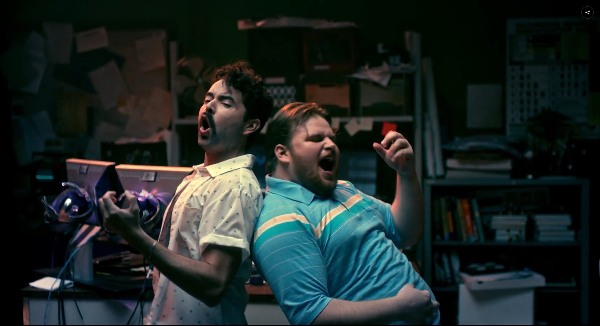 The Lab set on camera: two characters back to back mid-shout among paper-covered walls, metal shelving and stacked CRT monitors, lit teal and purple.