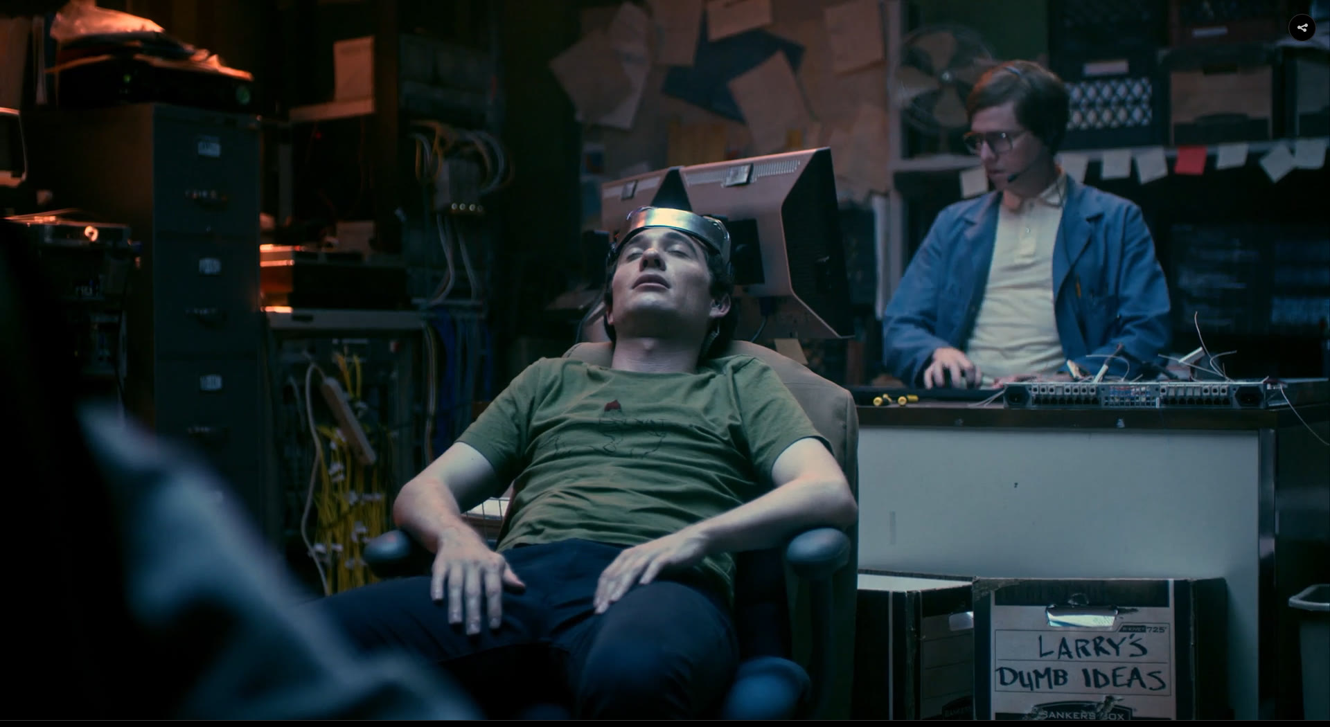 The Lab: a character reclined in the headset rig while another works a console behind him, surrounded by filing cabinets, cable racks and a bankers box labelled LARRY'S DUMB IDEAS.