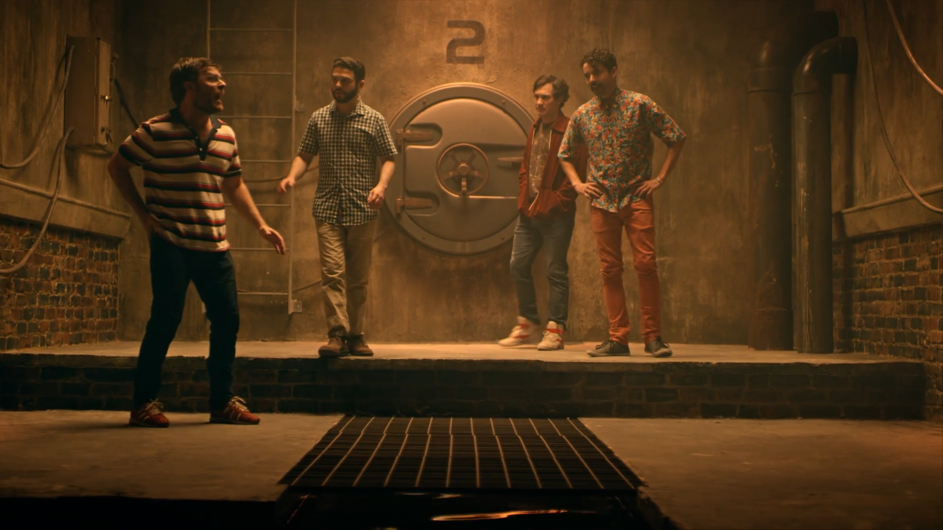 The Brainframe on camera: four cast members on the raised platform in front of the circular hatch door numbered two with its wheel lock, a ladder and standpipe against concrete-over-brick walls, and the steel floor grate open in the foreground.