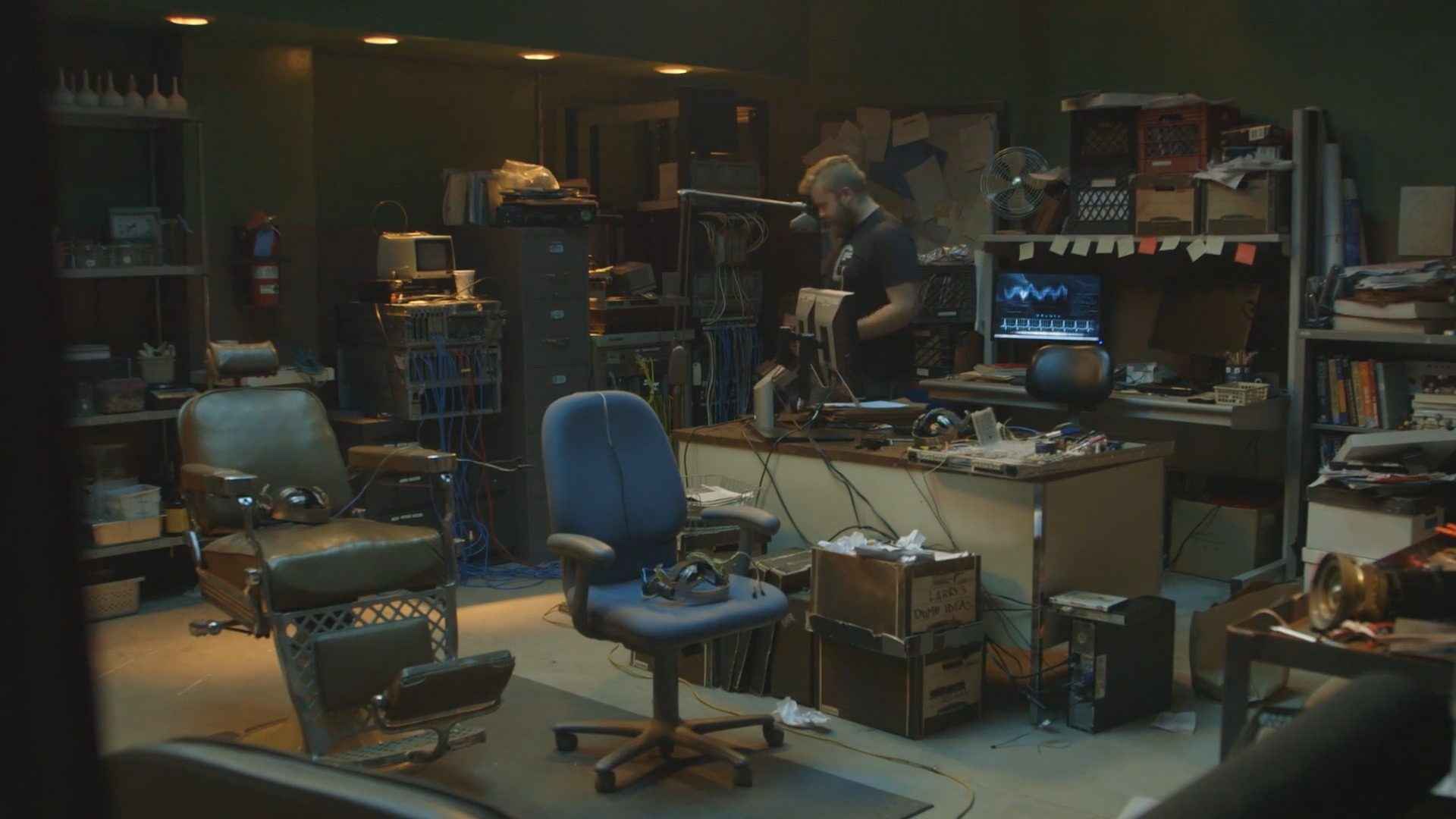 The Lab wide: a reclining leather chair and an office chair facing a bench of monitors and patched cable, with shelving, filing cabinets, a CRT, stacked boxes and a string of pennants filling the walls behind, and a figure working at the back.