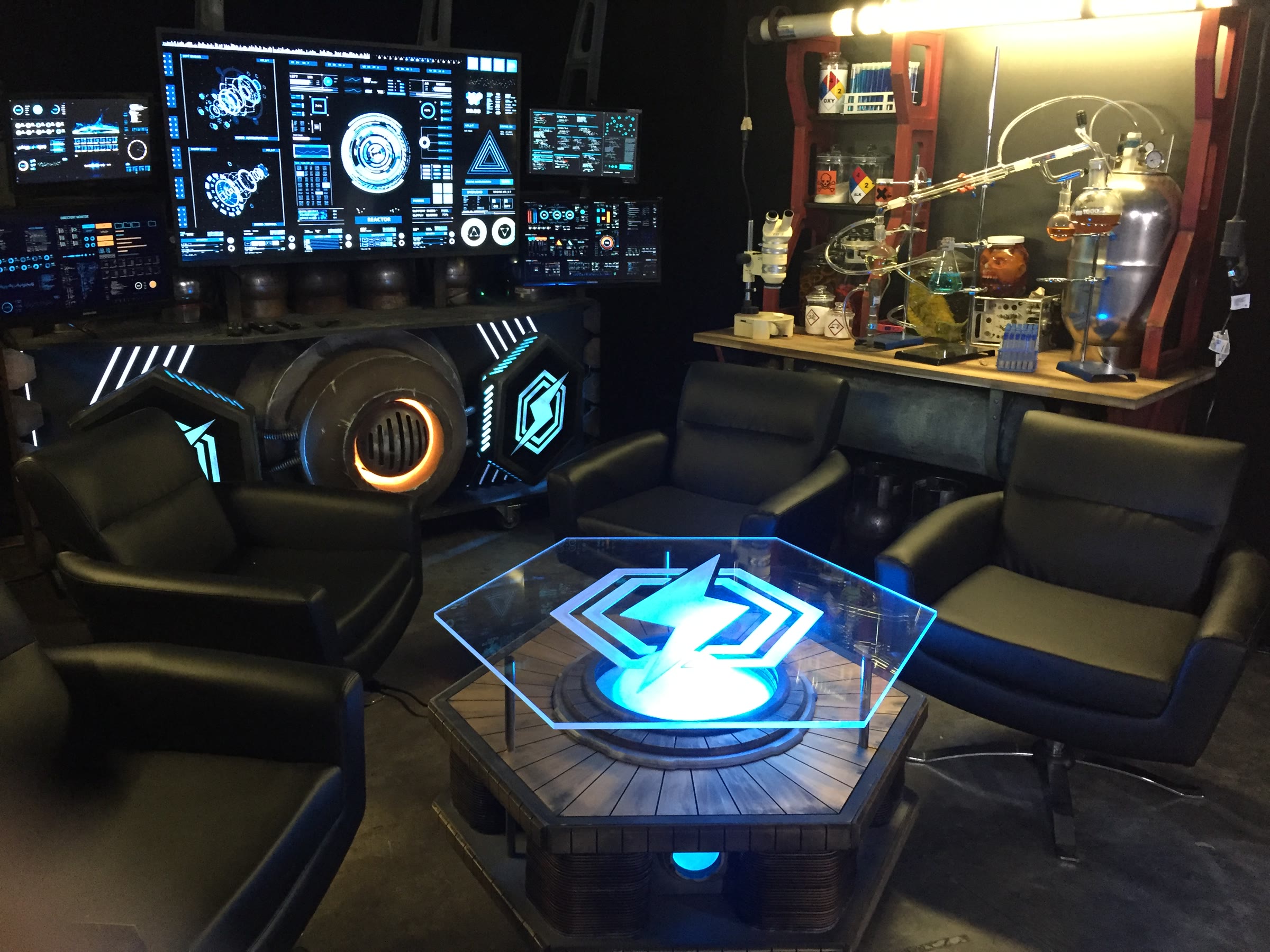 The assembled Death Battle set glowing on its first shoot: at left a control-panel backdrop with a bank of screens and a reactor-core cylinder, at right a lab bench of jars and glassware, and a hexagonal command table with a lit blue core ringed by four black chairs.
