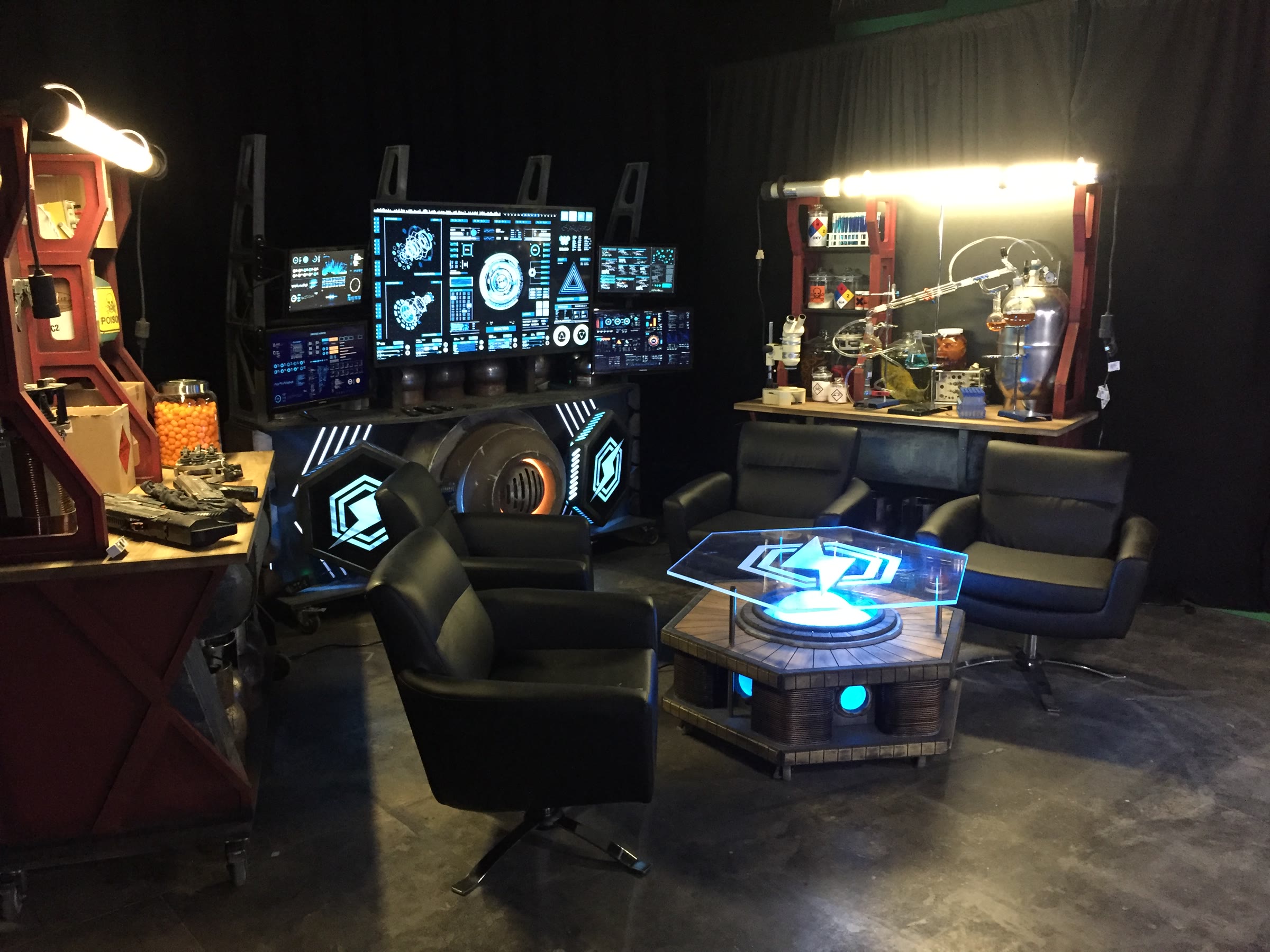 Wide view of all three set pieces plus the hex table: the armory bench of prop guns at left, the screen-and-reactor backdrop at center, and the red-framed lab tower at right, set up on a polished concrete floor before a black surround.