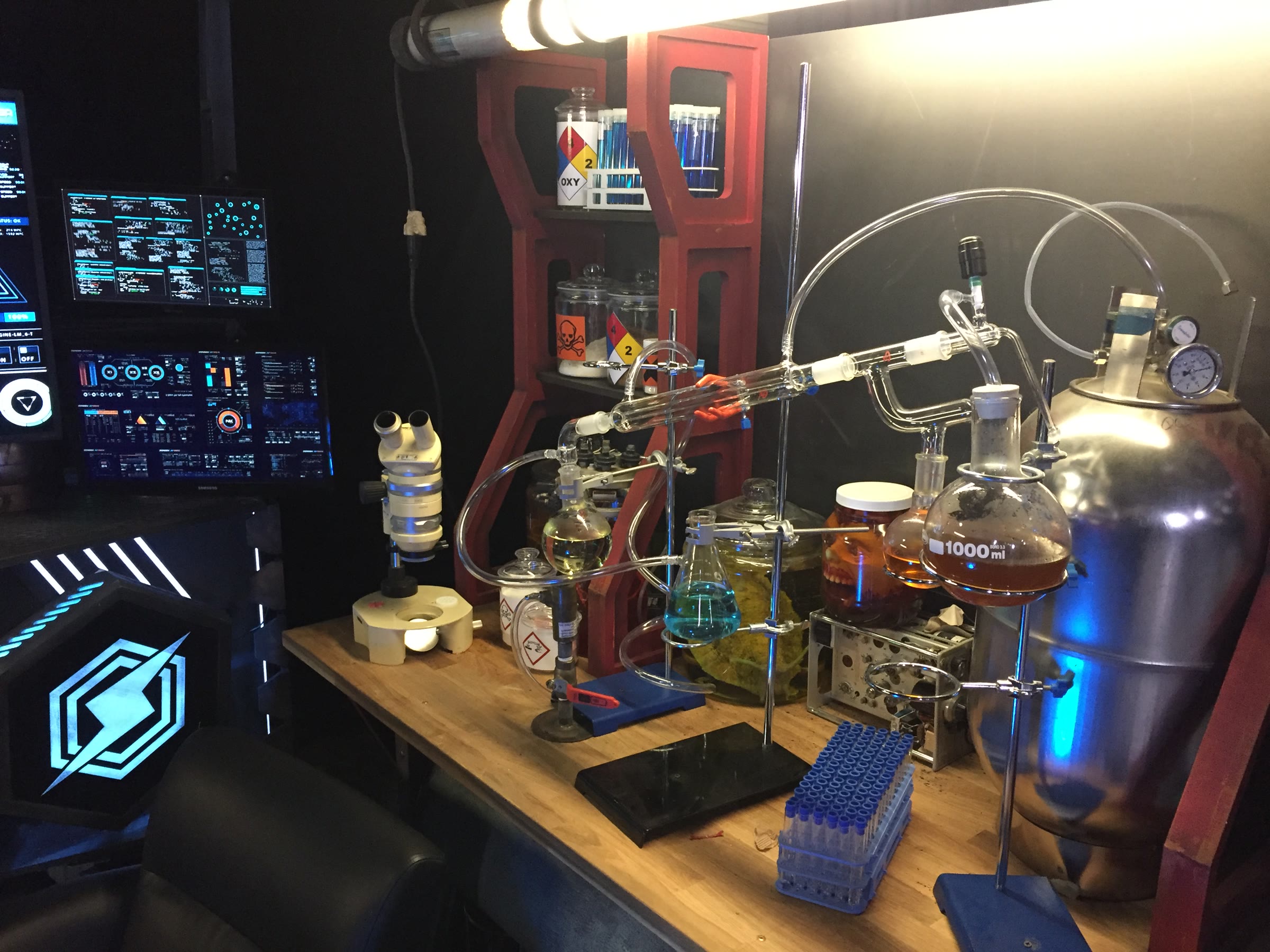 The lab bench in close-up: a microscope, a glass distillation train with colored liquids, a stainless pressure tank, hazmat-labeled jars, and a sculpted head suspended in an amber specimen jar.