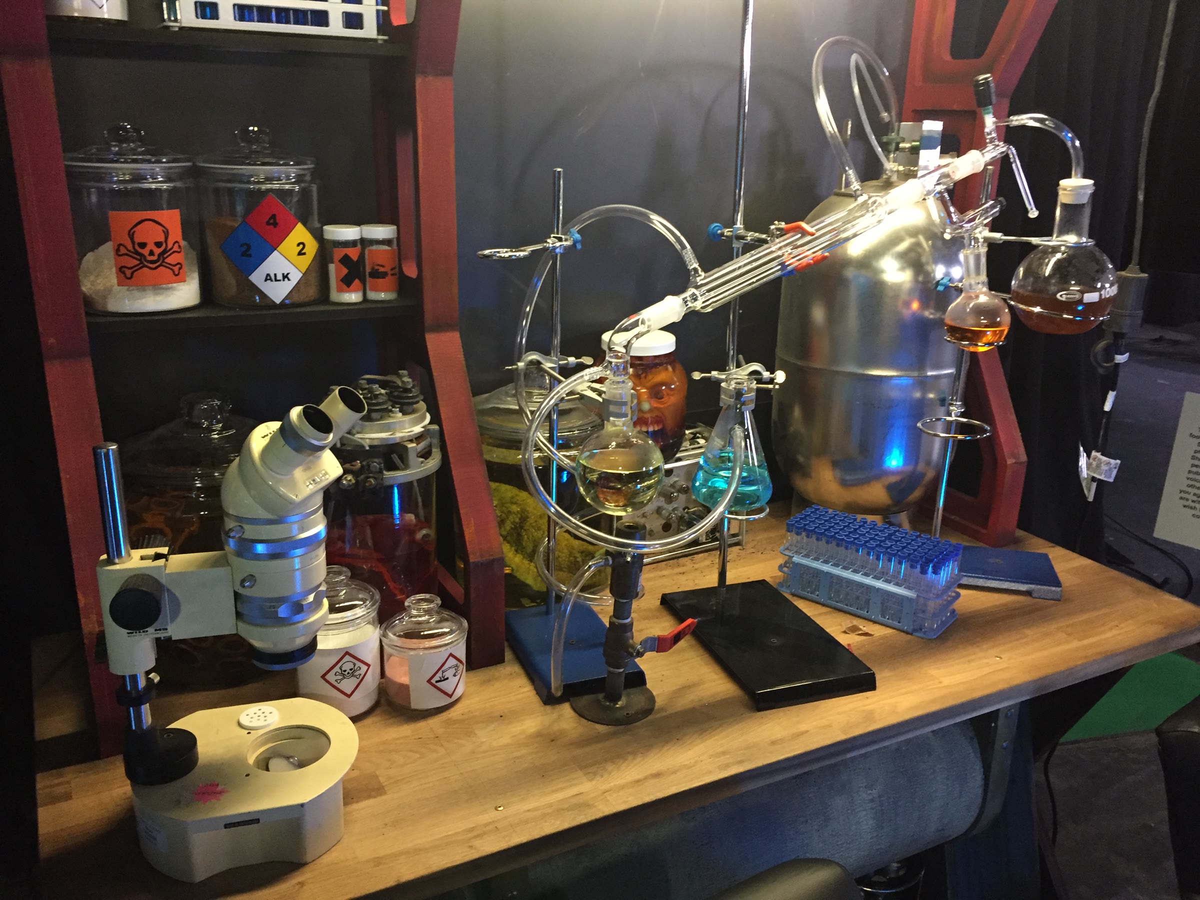 The built lab bench from the reverse angle: jars of powders with hazmat graphics, a stereo microscope, the head in an amber jar, a distillation train, and test-tube racks lit from below.