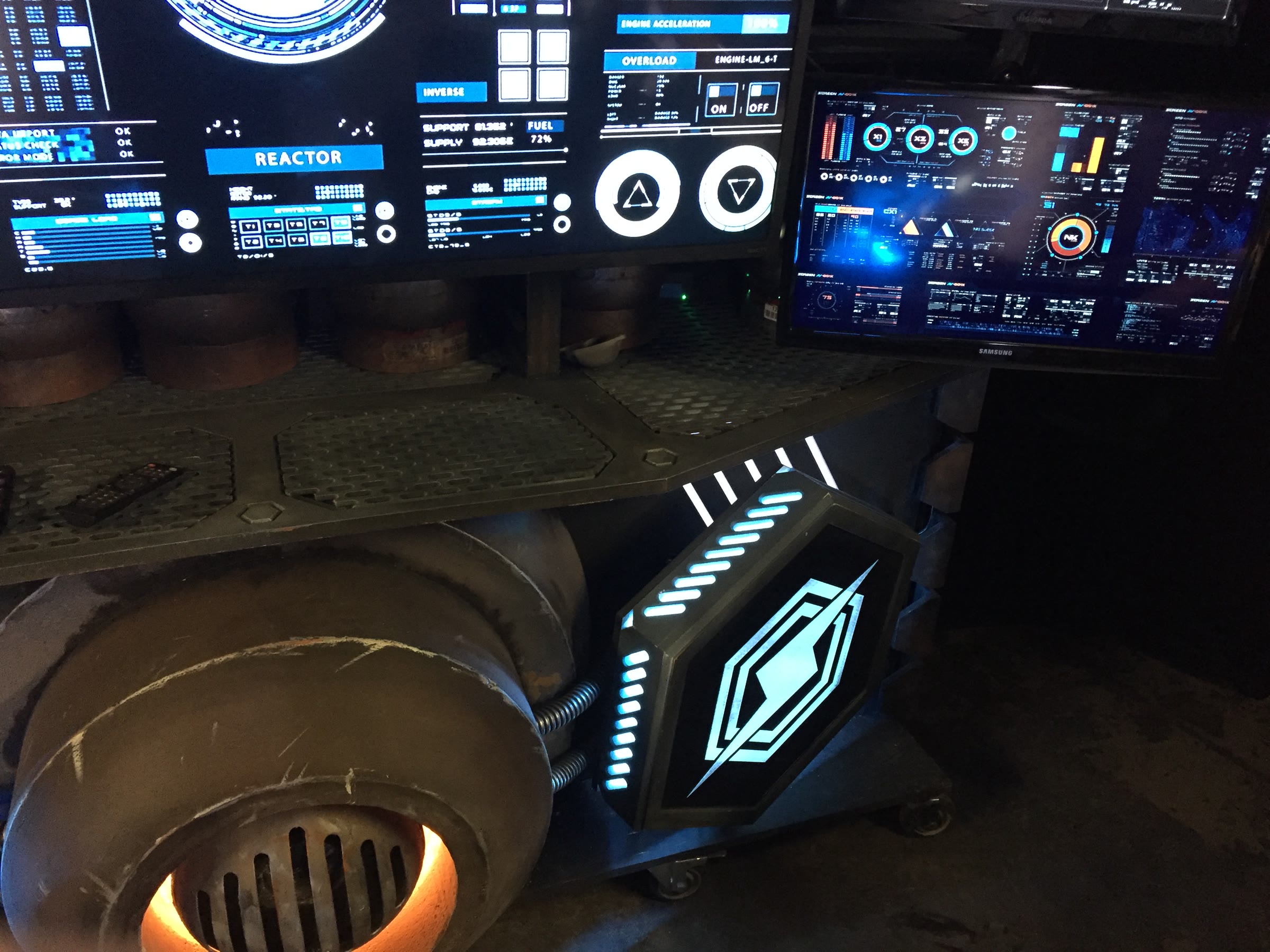 The built backdrop in detail: a bank of consumer screens running sci-fi interface graphics, a weathered reactor drum glowing orange from a slotted vent, hex-perforated deck plates, and casters under the steel base.