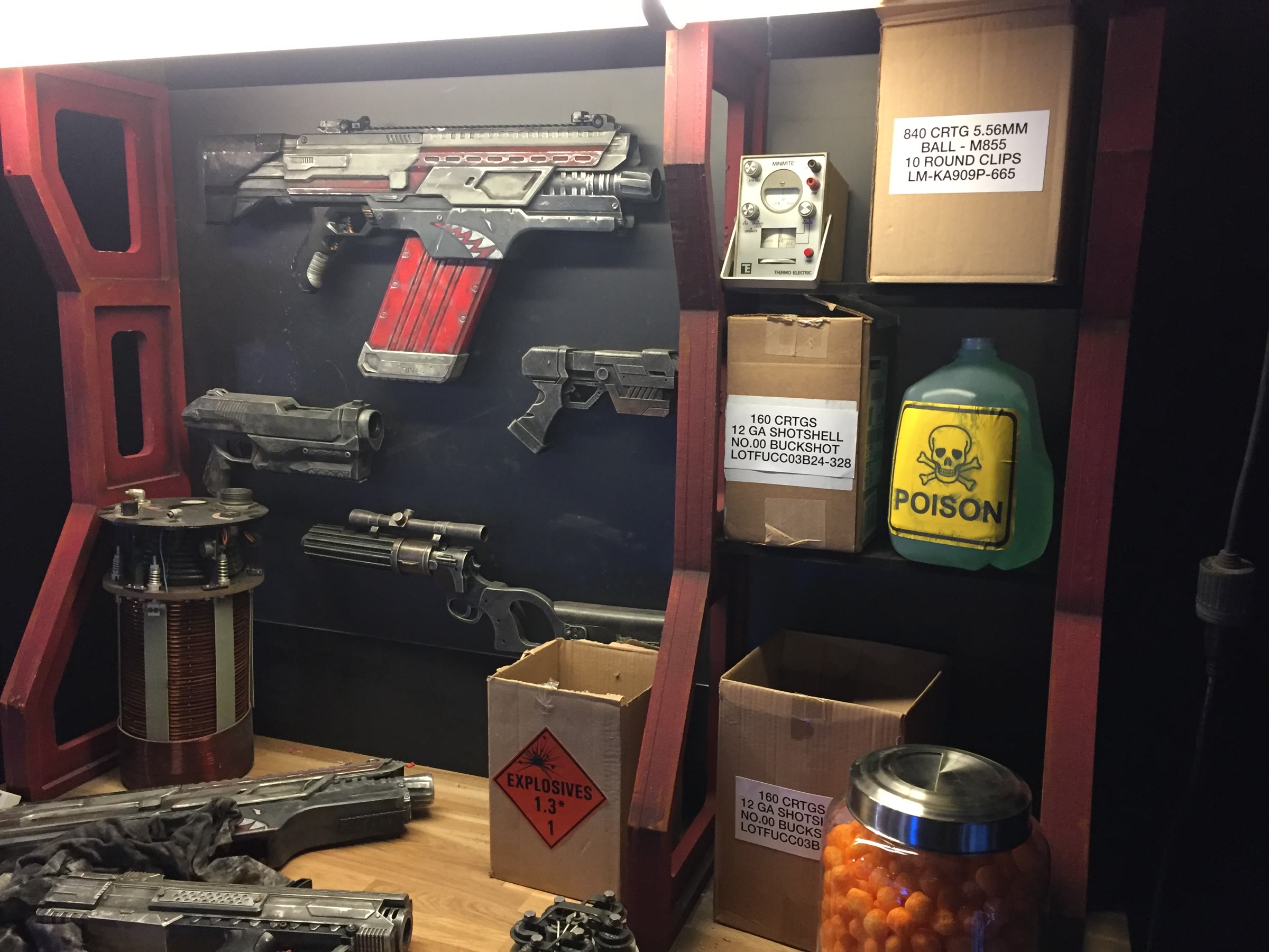 The armory wall: kitbashed Nerf guns weathered to gunmetal, a hero rifle with hand-painted shark teeth on a red magazine, pistols and a scoped long gun below, with printed ammo-crate labels.