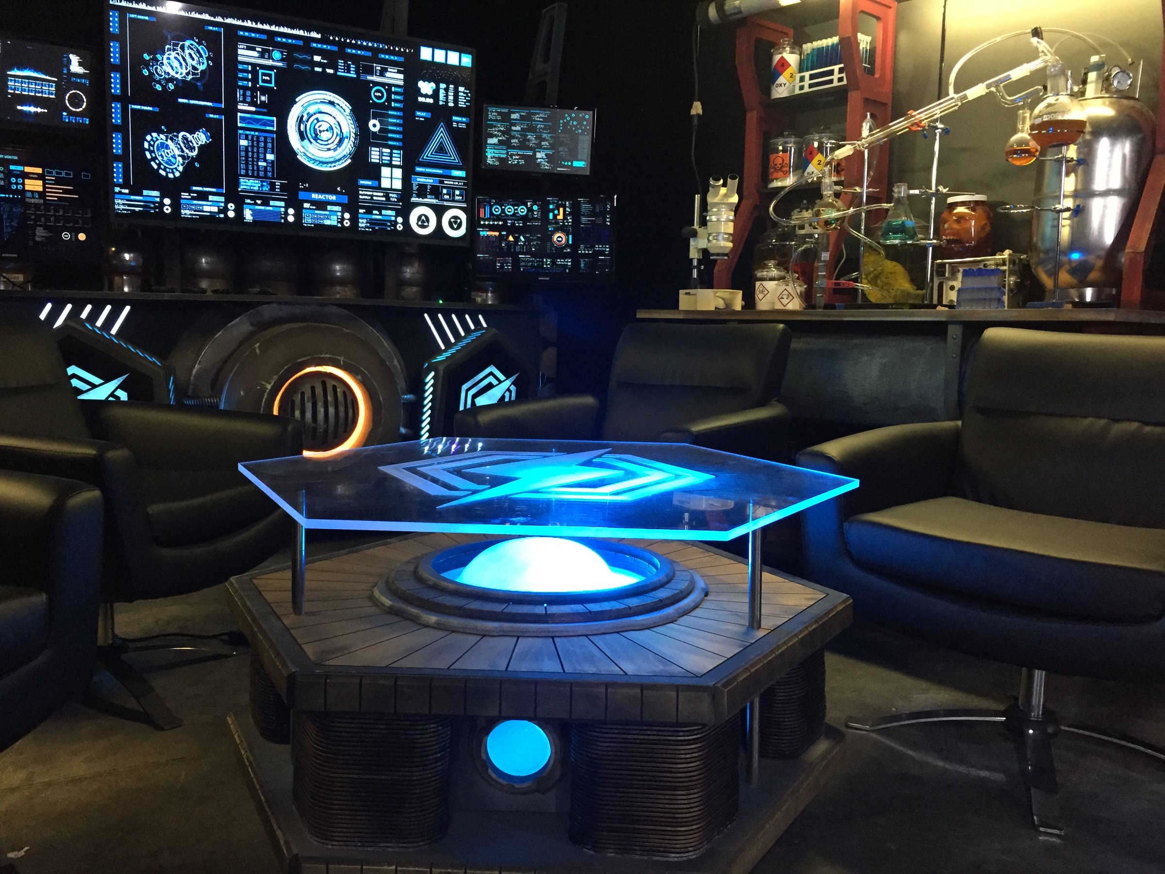 The hex table in a low beauty shot: an edge-lit clear-acrylic hexagon etched with the show's lightning-bolt logo, floating on steel standoffs above a blue-lit dome, on a base of ribbed copper-look heat-sink columns.