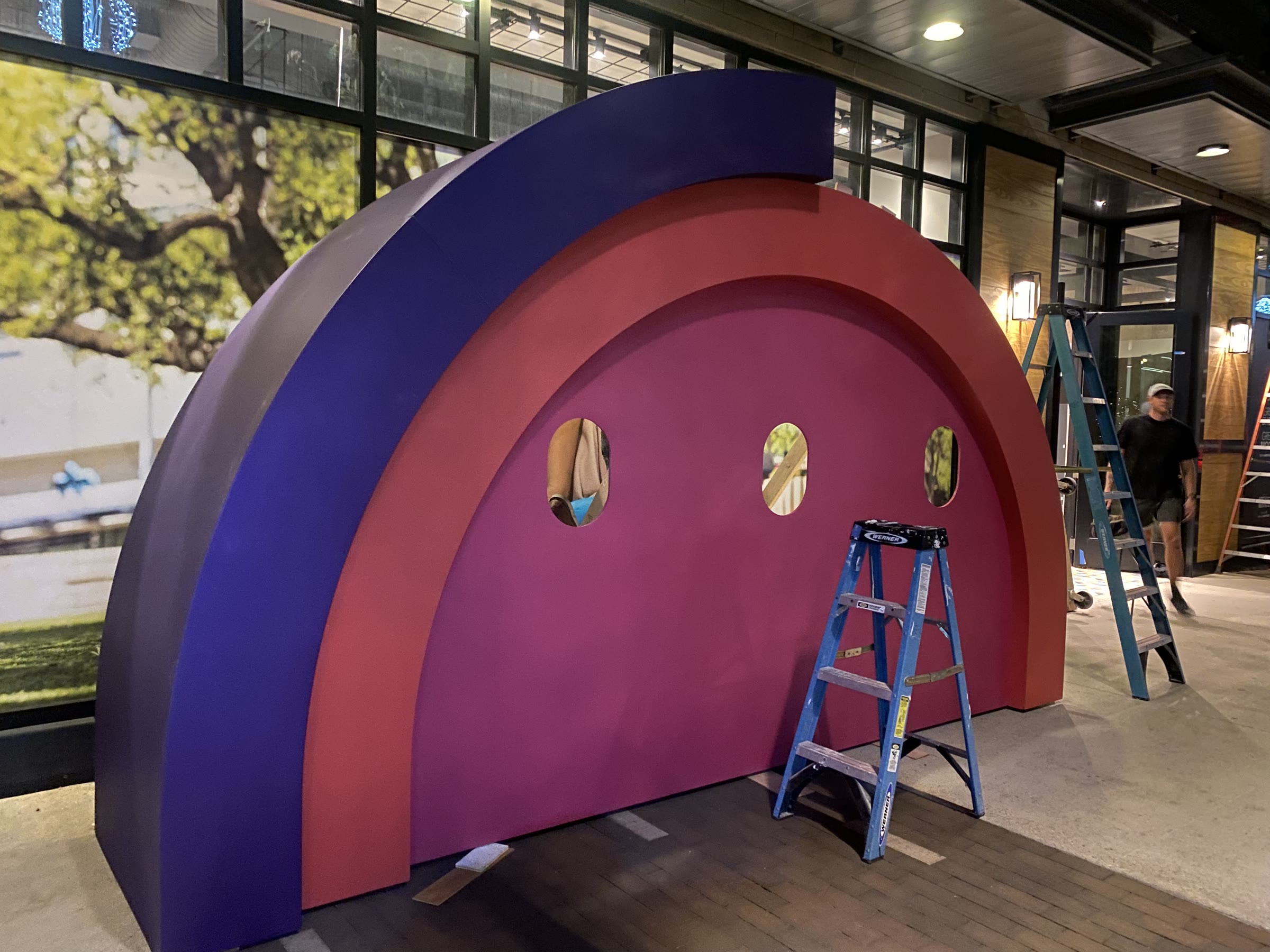 Install night on the Domain sidewalk: the rainbow arch stands assembled against a lit storefront, metallic purple outer band and red inner band around a magenta face with three empty oval porthole cutouts, two stepladders beside it and a crew member walking past.