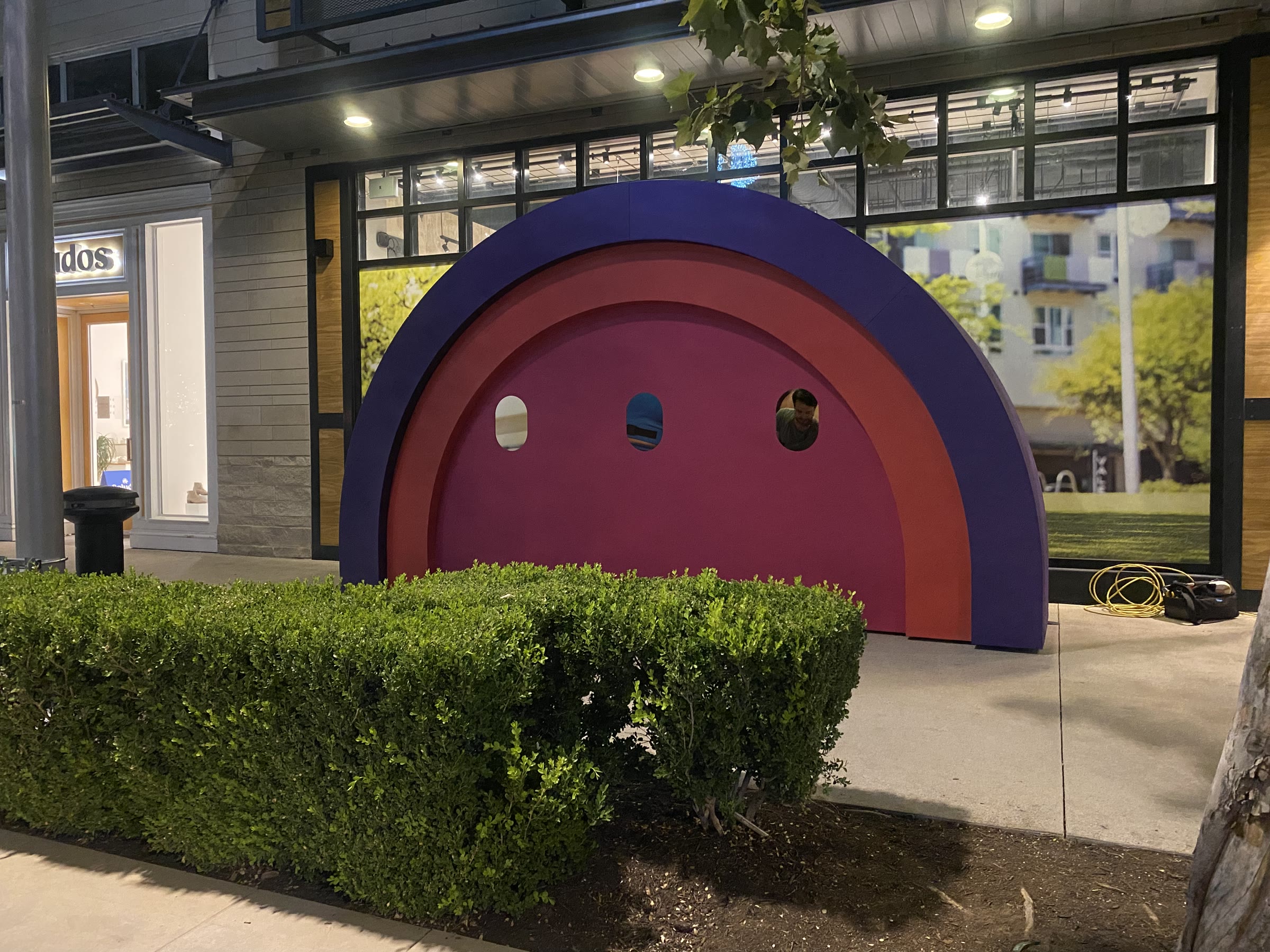 The assembled arch seen across a boxwood hedge at night, storefront glass lit behind it; a crew member looks out through the right-hand porthole, an extension cord coiled on the pavement beside the base.