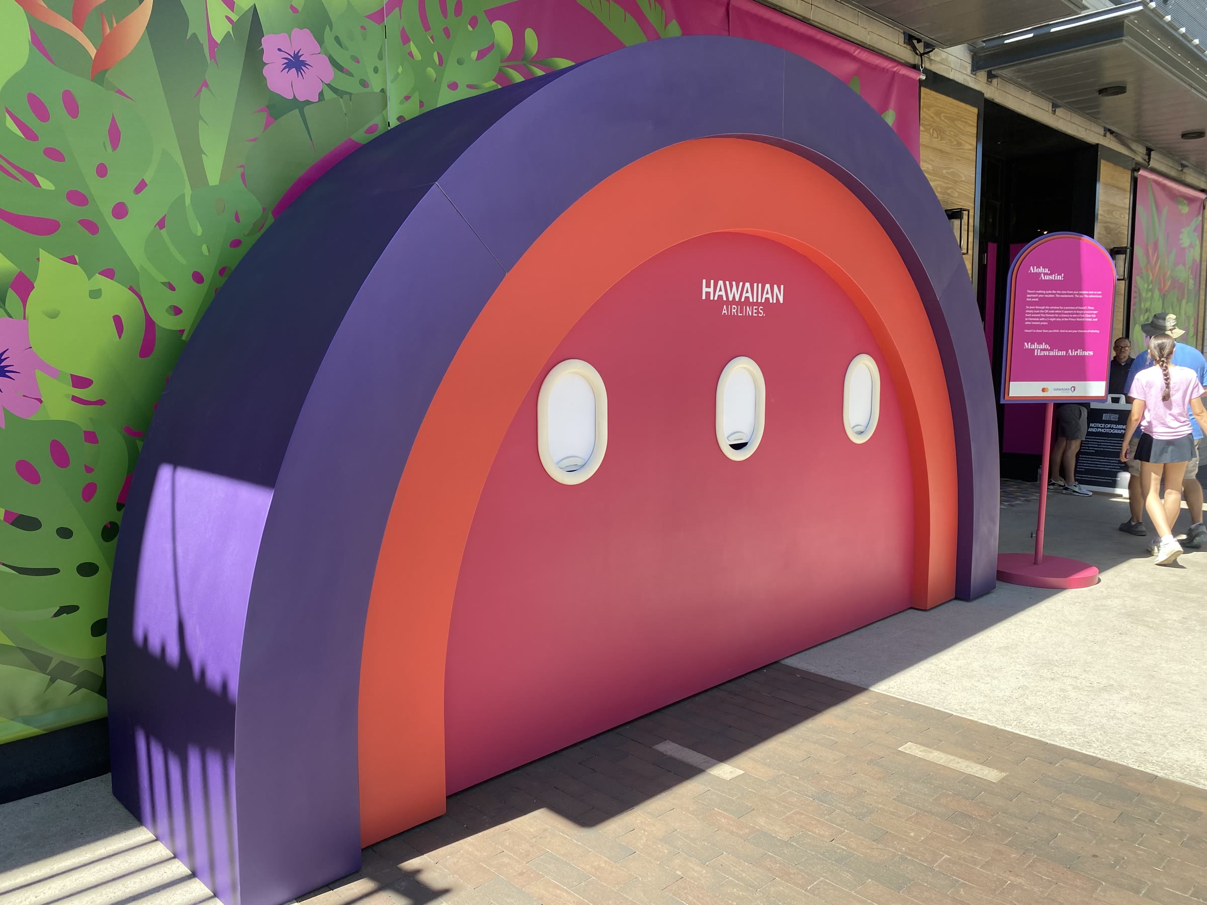 The finished activation on the Domain sidewalk: a walk-up half-dome arch in concentric bands of purple, coral, and magenta, with the HAWAIIAN AIRLINES wordmark and three oval airplane windows on the pink inner face; a tropical botanical mural on the banner wall behind it.