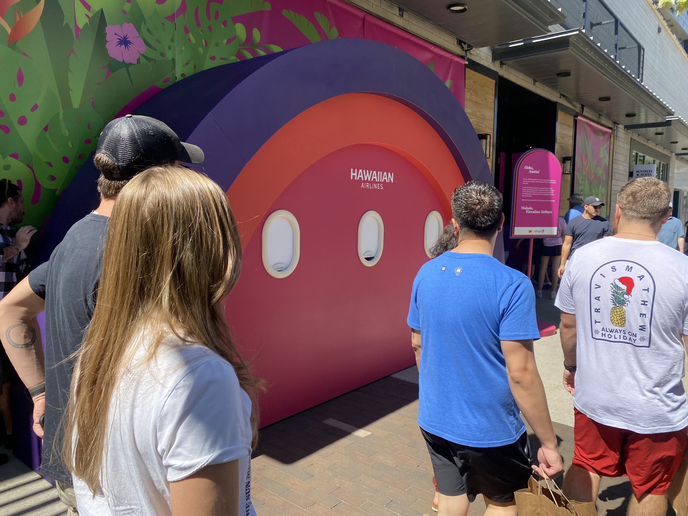 The arch on the sidewalk with passers-by walking up to it: concentric purple, coral, and magenta bands, the HAWAIIAN AIRLINES wordmark and three oval windows on the pink face, the tropical mural banner behind.