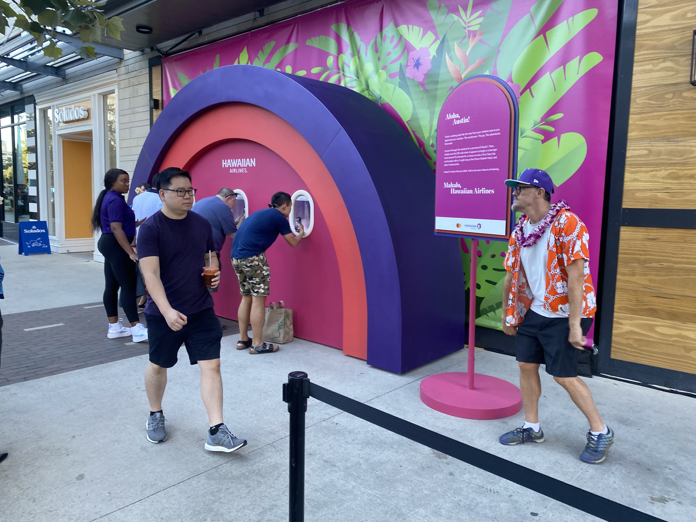 A side view of the arch showing its depth as a half-tunnel; several people bend to peer into the windows, one holding a phone up to the glass, with the botanical mural banner and a storefront behind.