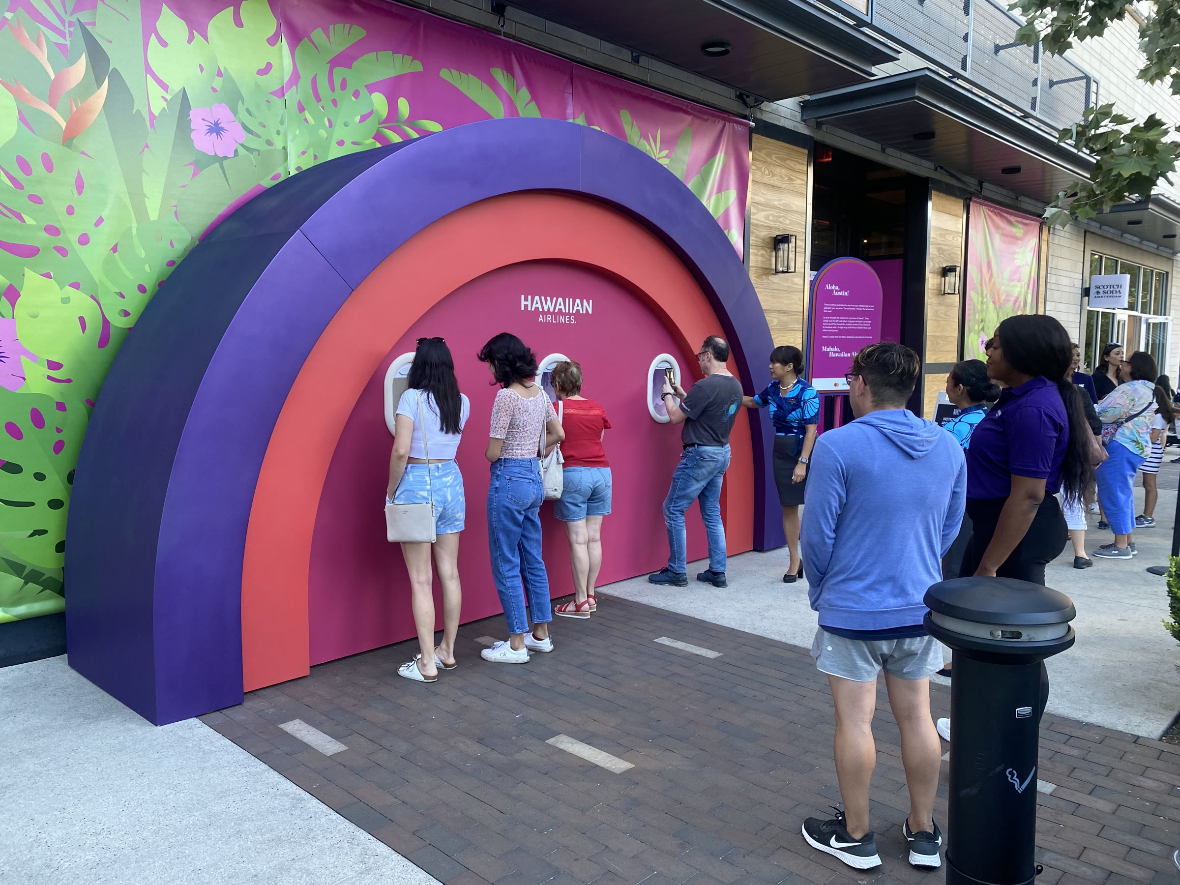 Evening at the Domain: a line of visitors lean in to look through the three oval windows on the pink face of the arch, a Hawaiian Airlines staffer in an aloha-print shirt beside them, a retail storefront to the right.