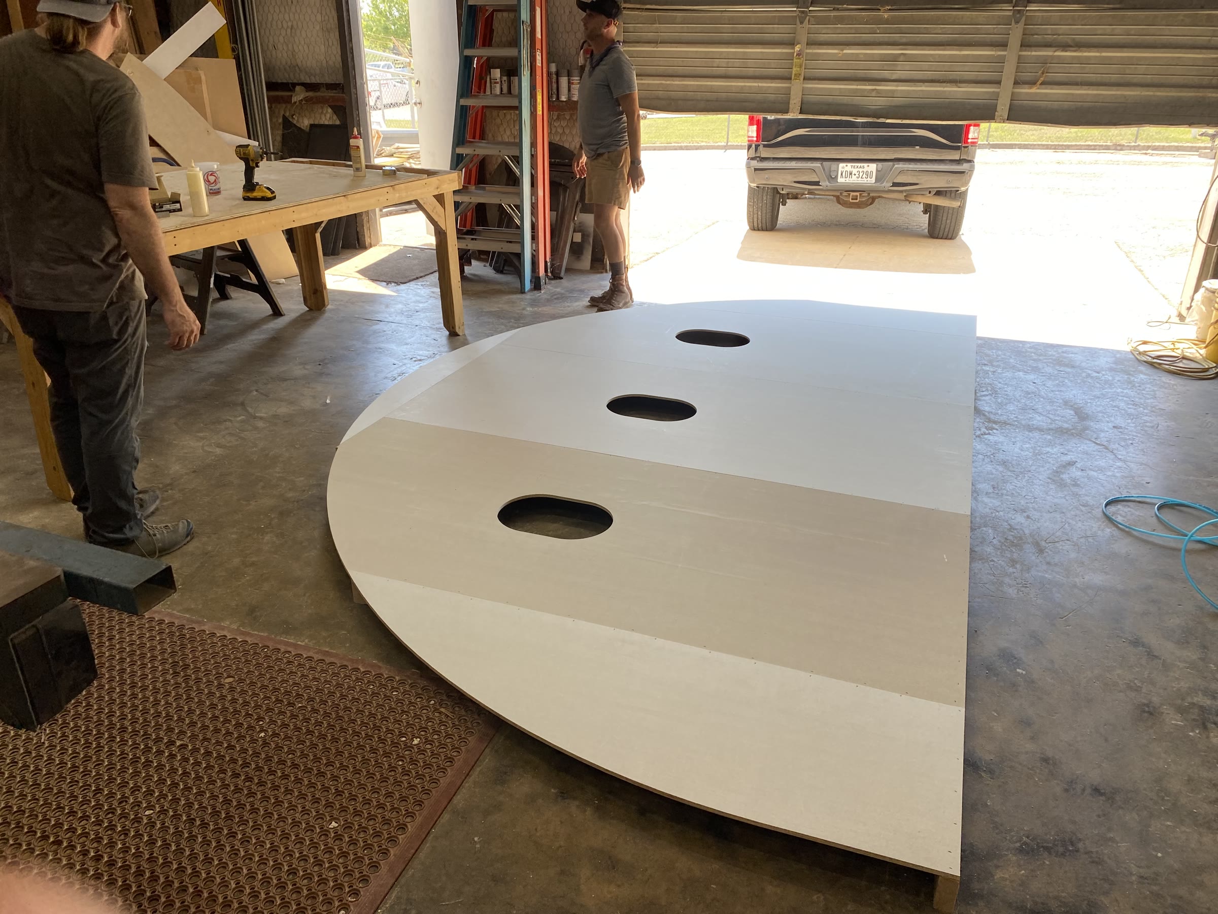 In the shop: a large semicircular plywood panel primed white lies flat on the concrete floor with three oval airplane-window openings cut into it, two crew members standing over it, a pickup backed up to the open roll-up door beyond.