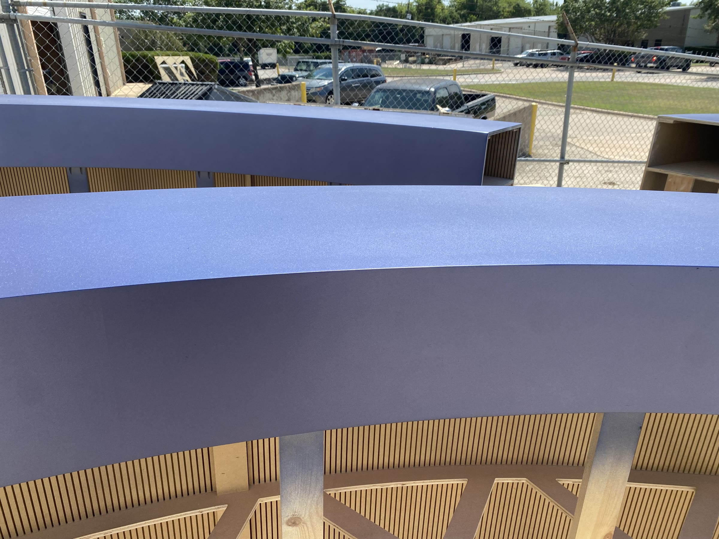 A curved arch segment finished in metallic purple, its wooden rib framework visible along the underside, standing in the fenced shop yard under a bright sky.
