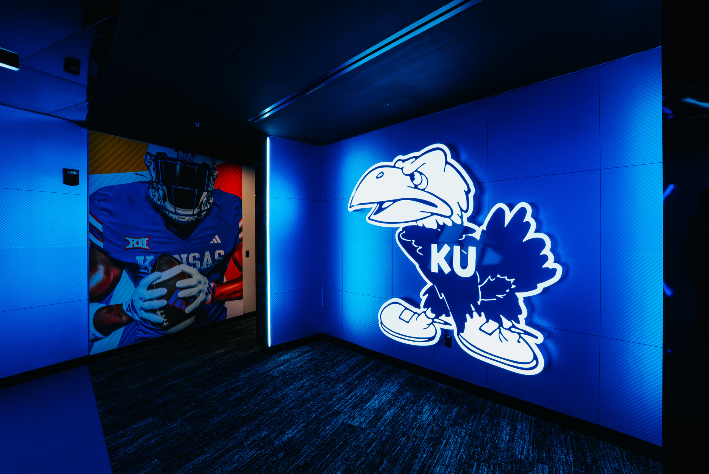 Gear display entry corner: the backlit vintage walking Jayhawk on the divider wall beside the player-photo transition mural striped in orange and red.