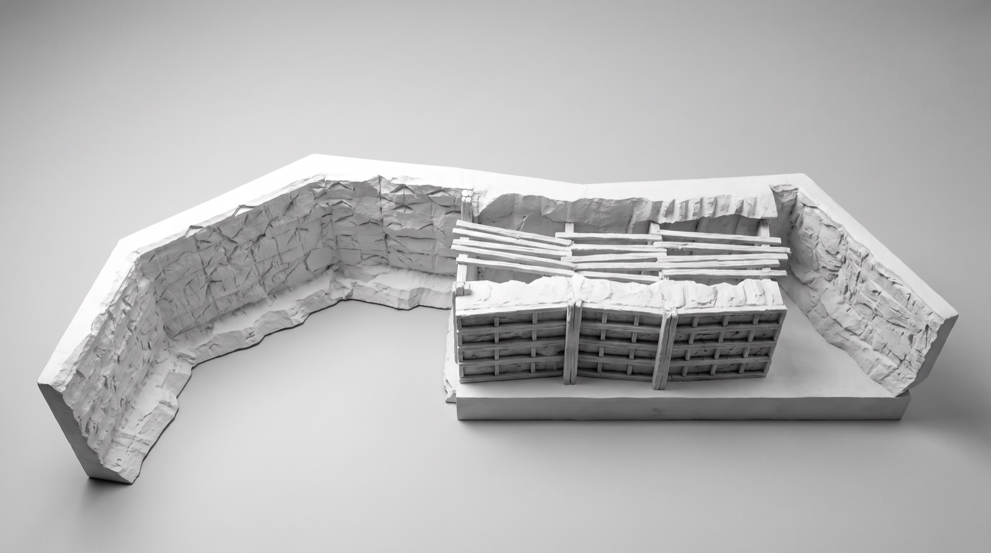 Untextured clay render of the set model seen from above: a long serpentine run of sculpted rock wall sweeping in from the left, and at right a timber-framed structure of stacked rails and posts sitting on a raised platform deck.