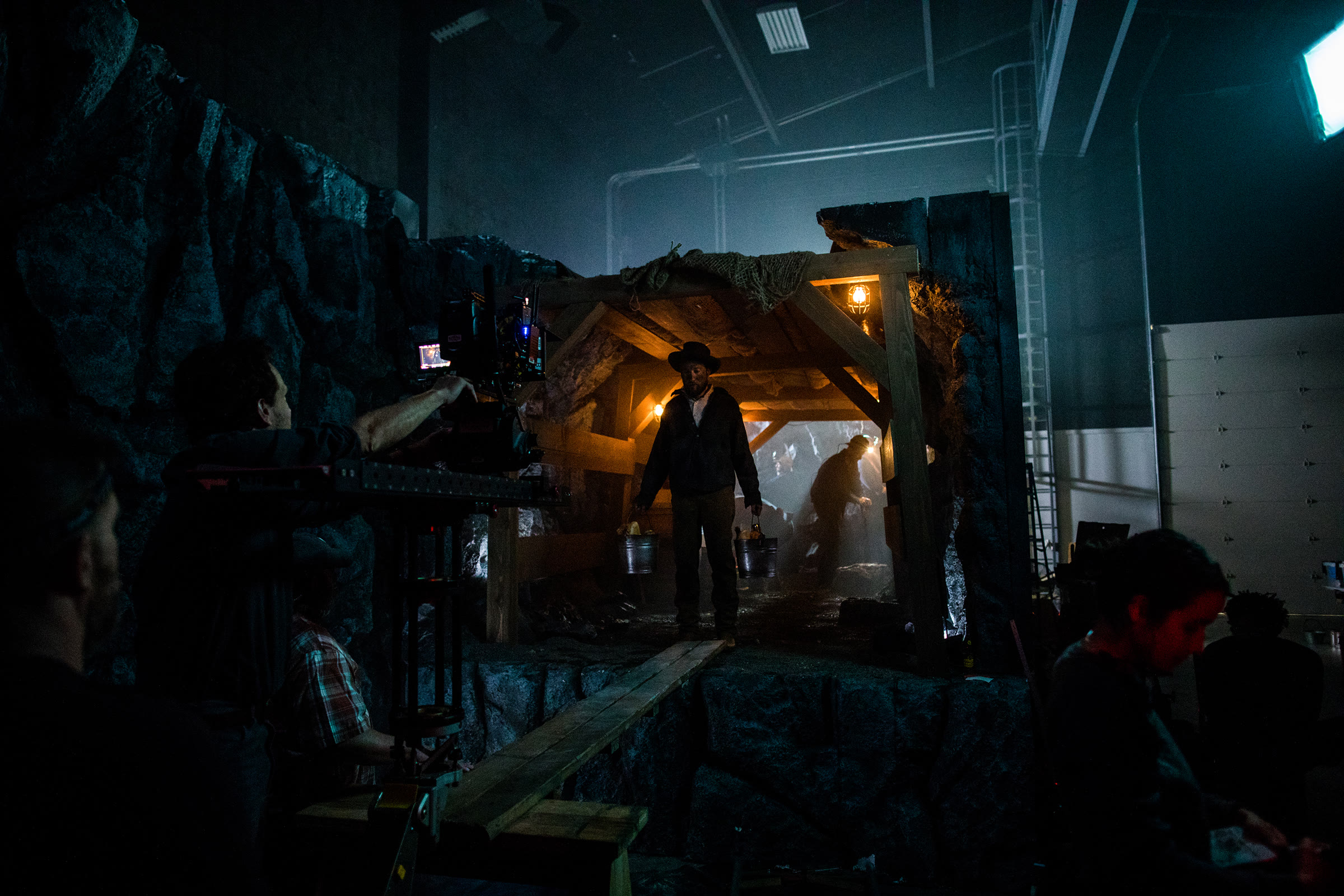 Behind the scenes on set: a section of the timber-framed tunnel with raked, off-square beams and burlap over the top, a costumed miner inside carrying buckets, with the soundstage ceiling grid, a ladder and stage walls deliberately visible around the set.