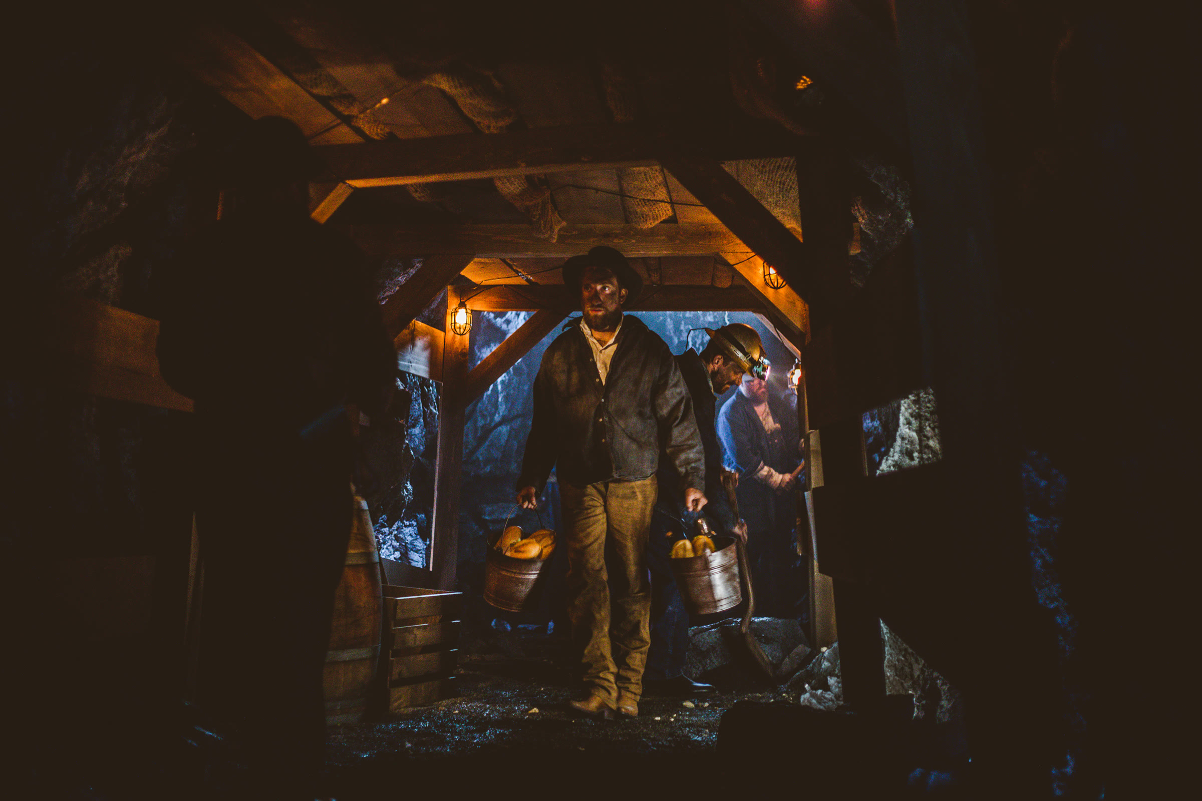 Inside the built tunnel, mid-take: a costumed miner in a hat carries two metal ore buckets down the shaft, a second miner with a lit helmet lamp and a shovel behind him, a barrel and crate to the left, caged practical lamps on the timber, rubble underfoot.