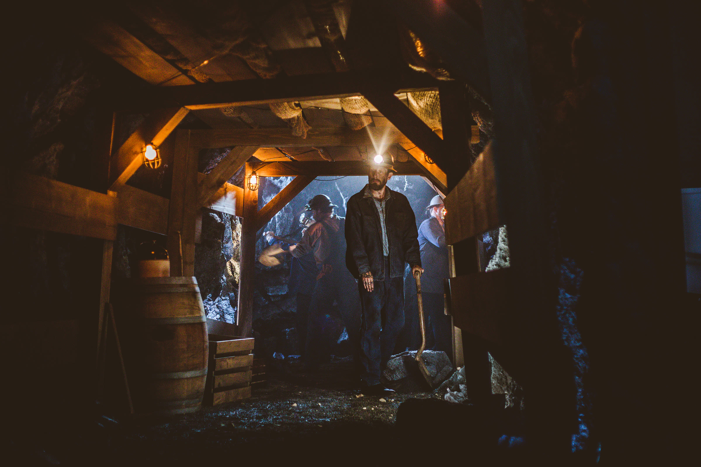 Deeper in the built tunnel: a miner with a lit headlamp and shovel at centre and two more behind in blue haze, surrounded by a rich dressing layer — barrel, crate, a ceramic jug on a rail, burlap netting between the roof timbers, and a rubble floor.