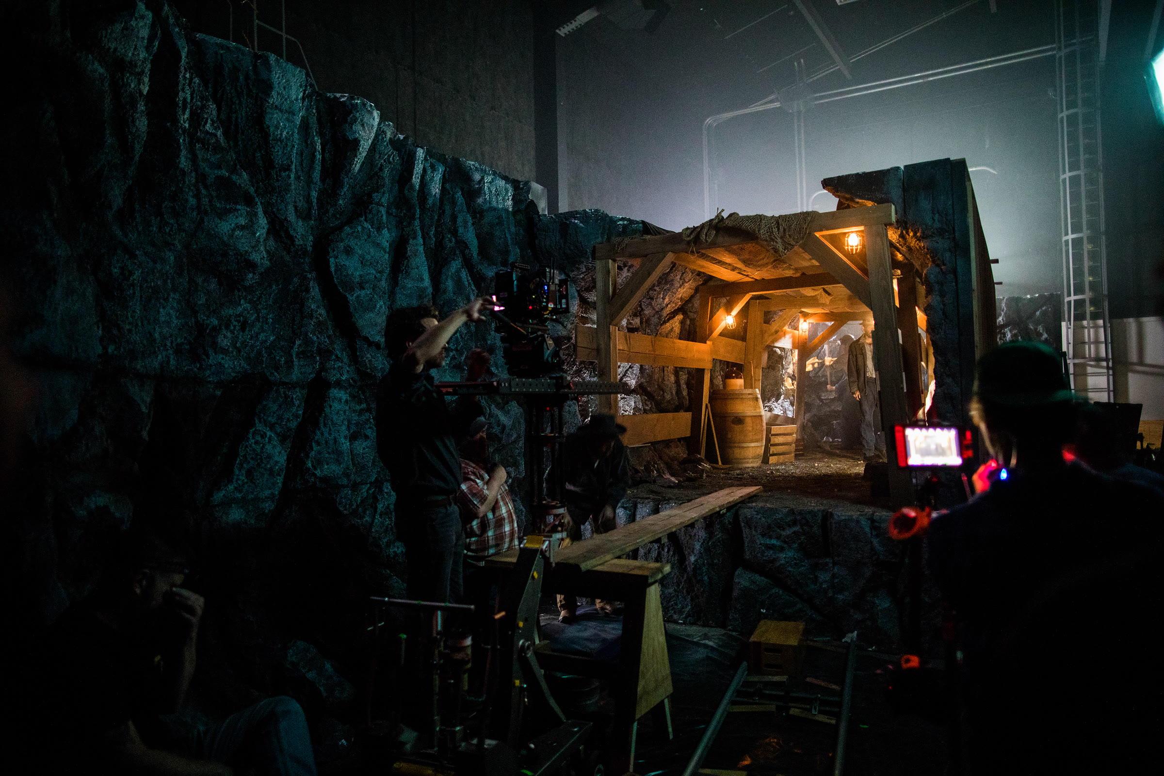 Behind the scenes on the soundstage: a blue-lit carved-rock wall with a film crew and camera at its foot on the left, and on the right a raised platform holding a warm-lit, timber-framed mine-tunnel mouth, lit practicals hanging from its beams.