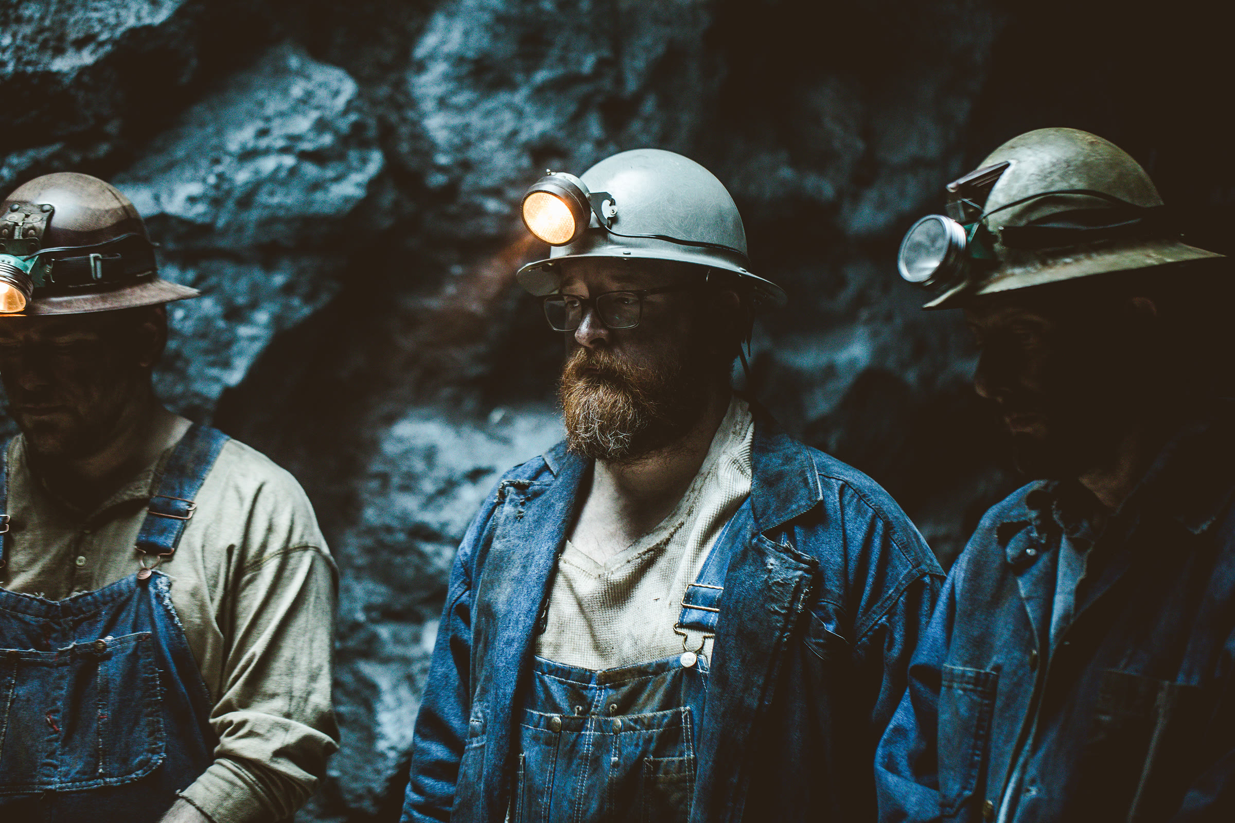 Tight three-shot of costumed miners against the carved rock wall, antique headlamps mounted on all three hard hats; the centre miner's lamp is lit.