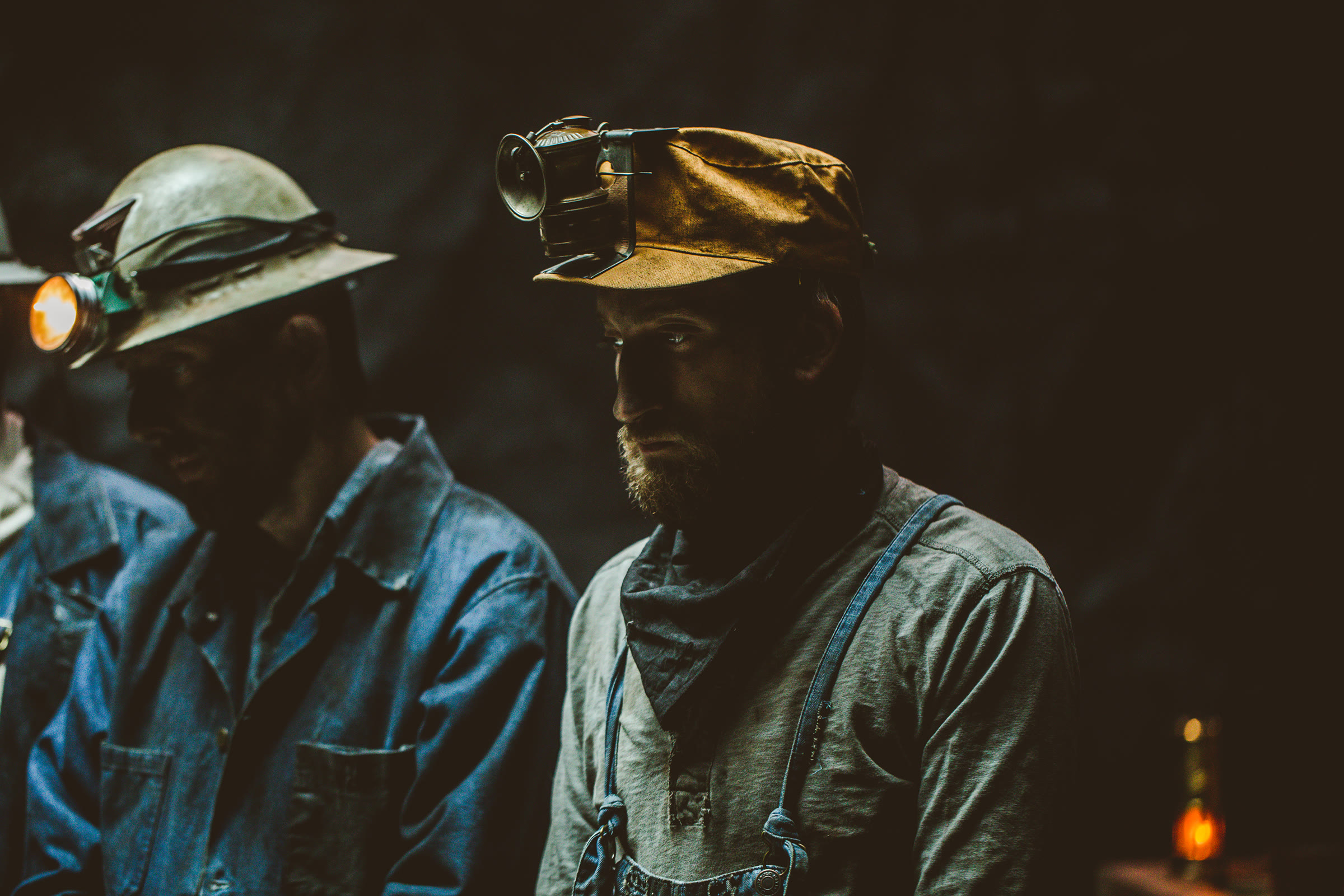 Two costumed miners side by side: the left in a metal hard hat with a lit lamp, the right in a soft canvas cap with a different carbide-style reflector lamp, showing the variety of period lamps across characters.