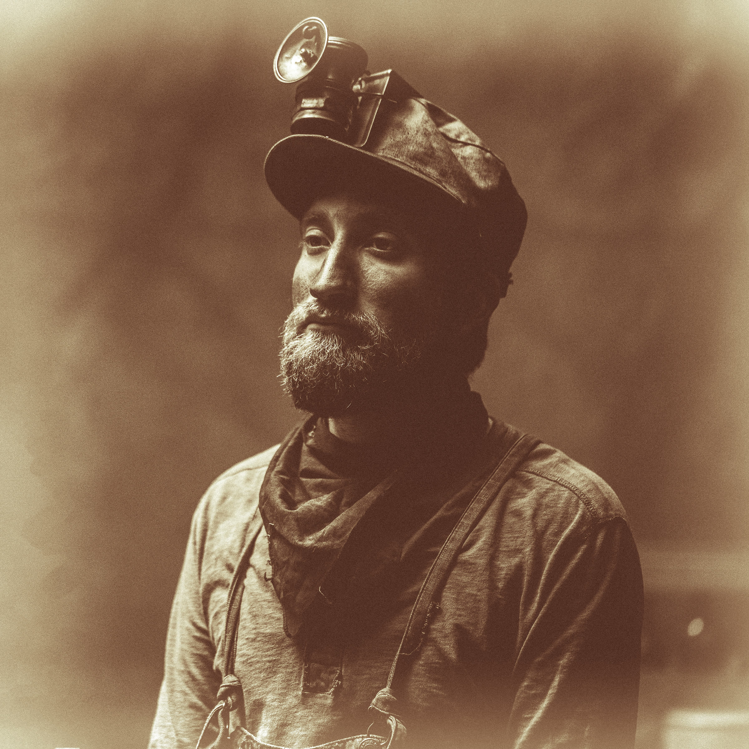 A single production still of a soft-cap miner retouched into a period photograph: sepia tone, a plate-style vignette fade and film grain turn the shot into an in-world 1800s miner's portrait.