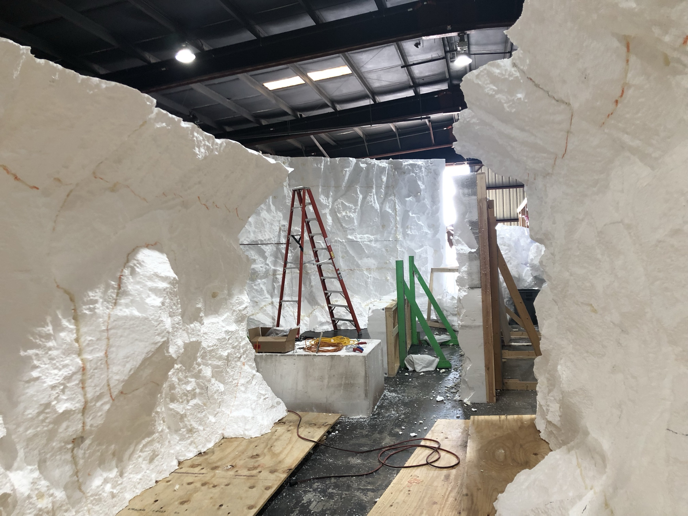 Inside the tunnel mid-build at the shop: carved styrofoam walls still raw white, a ladder against the far wall, plywood sheets and foam scraps on the floor.