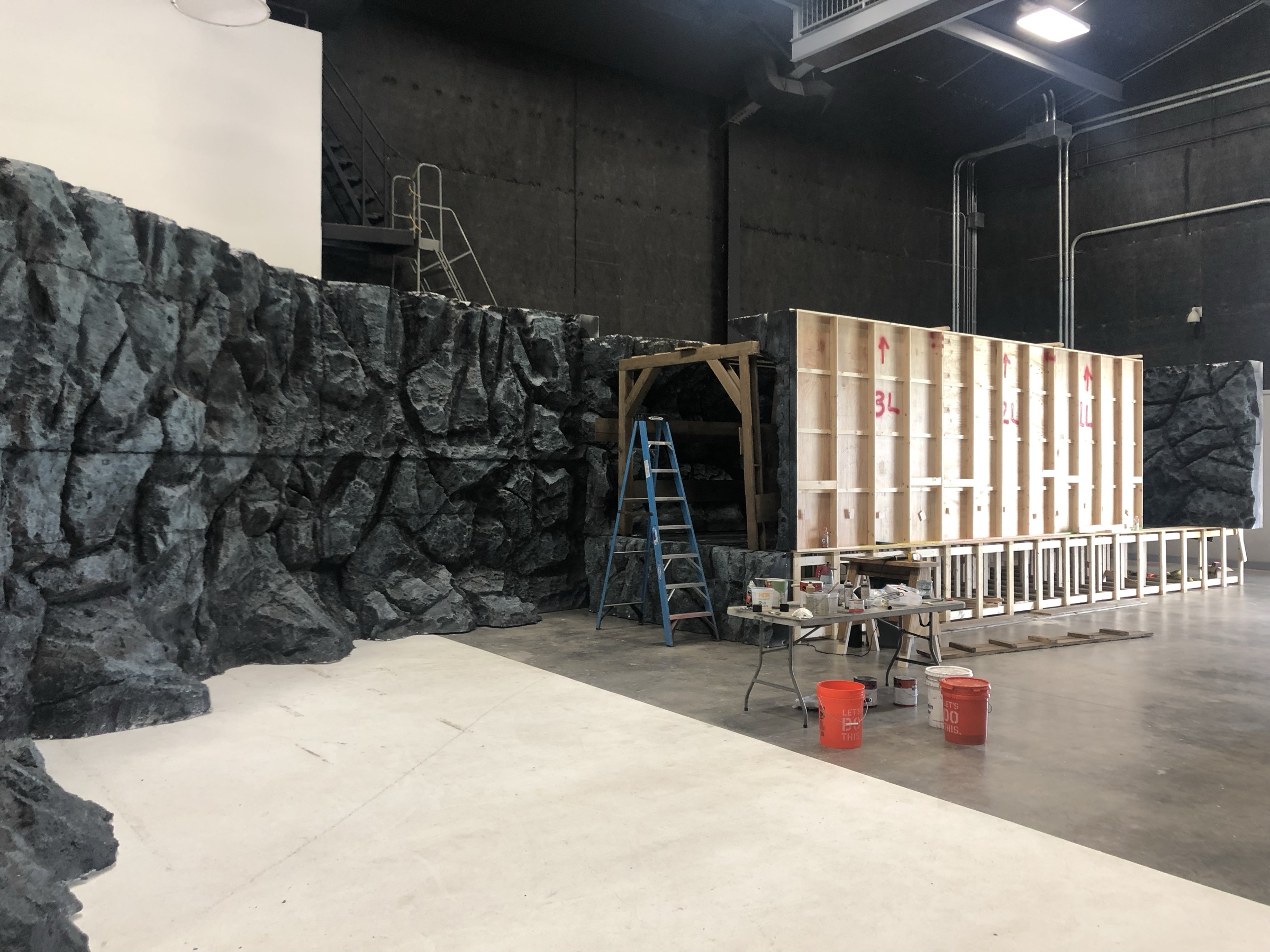 The carved rock walls standing on the soundstage mid-assembly: painted stone faces at left, the raw plywood backs of two flats still exposed at right, a work table and paint buckets in the foreground.