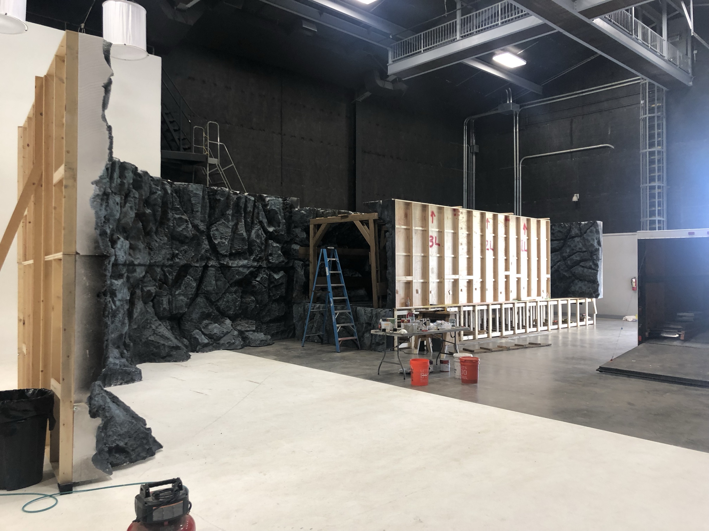 Wide view of the soundstage during assembly: painted rock sections standing at left, the exposed plywood backs of unpainted flats at right, a paint table and buckets between them.
