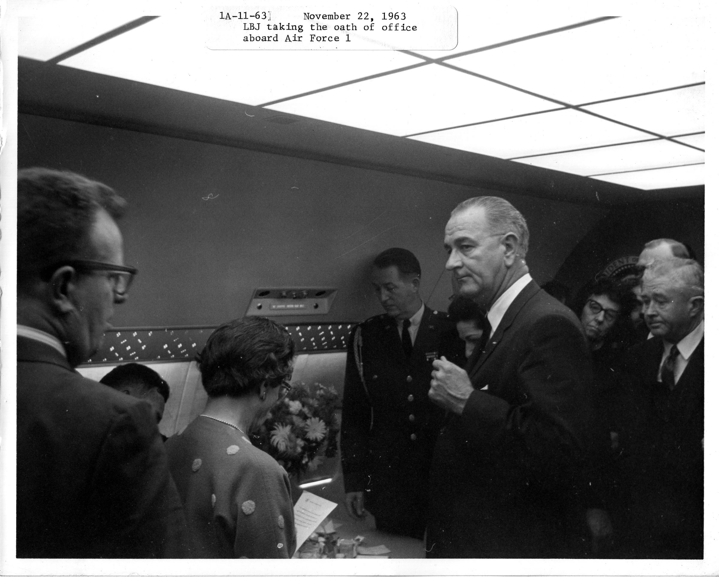 An archive photograph stamped November 22, 1963: Lyndon Johnson taking the oath of office aboard Air Force One, surrounded by officials under the cabin's luminous ceiling grid and perforated speaker band.