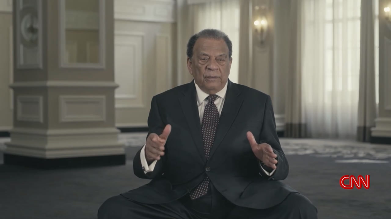 A filmed interview: an interviewee in a dark suit speaking to camera in a grand paneled room, hands raised mid-thought.