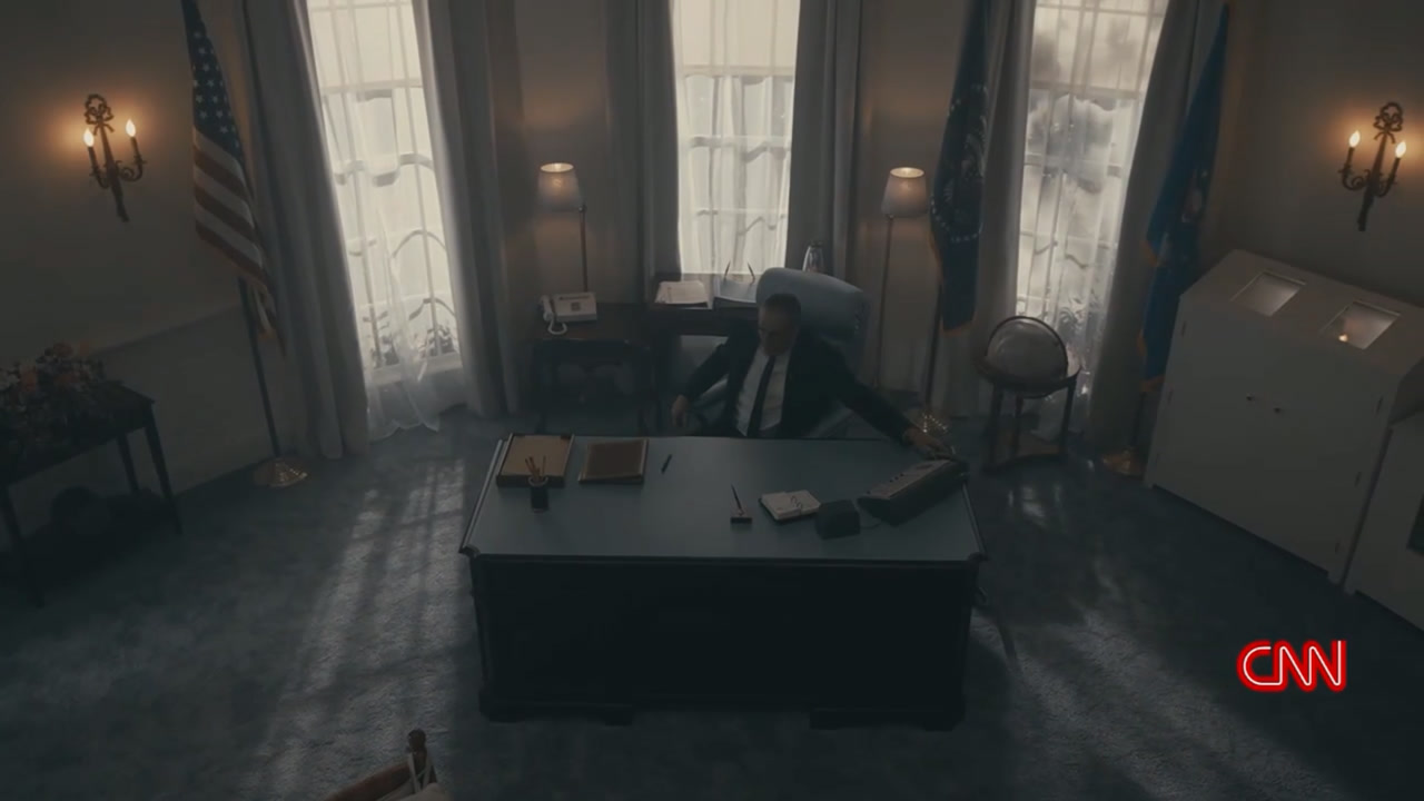 A reenactment frame: the actor at the Oval Office desk in low light on the finished set, flags and lit windows behind him.