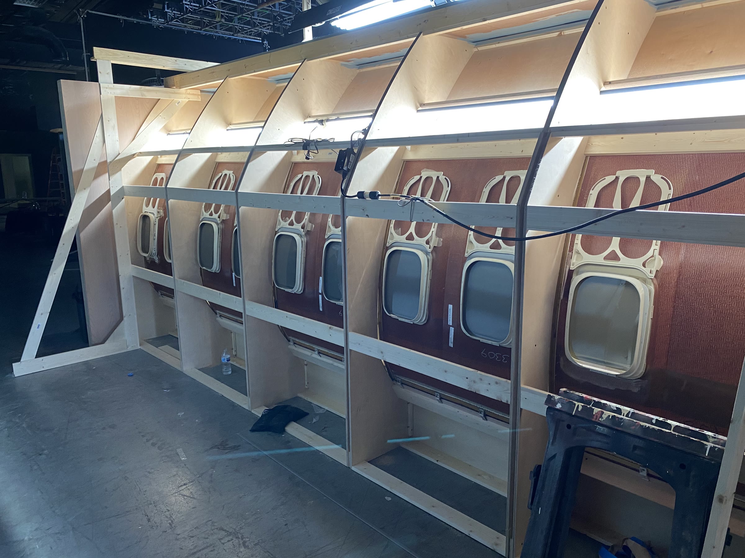 The fuselage wall seen from behind the set: stud-and-plywood framing carrying real salvaged airliner sidewall panels with molded cream double-porthole surrounds, and fluorescent worklights rigged above to daylight the portholes from outside.