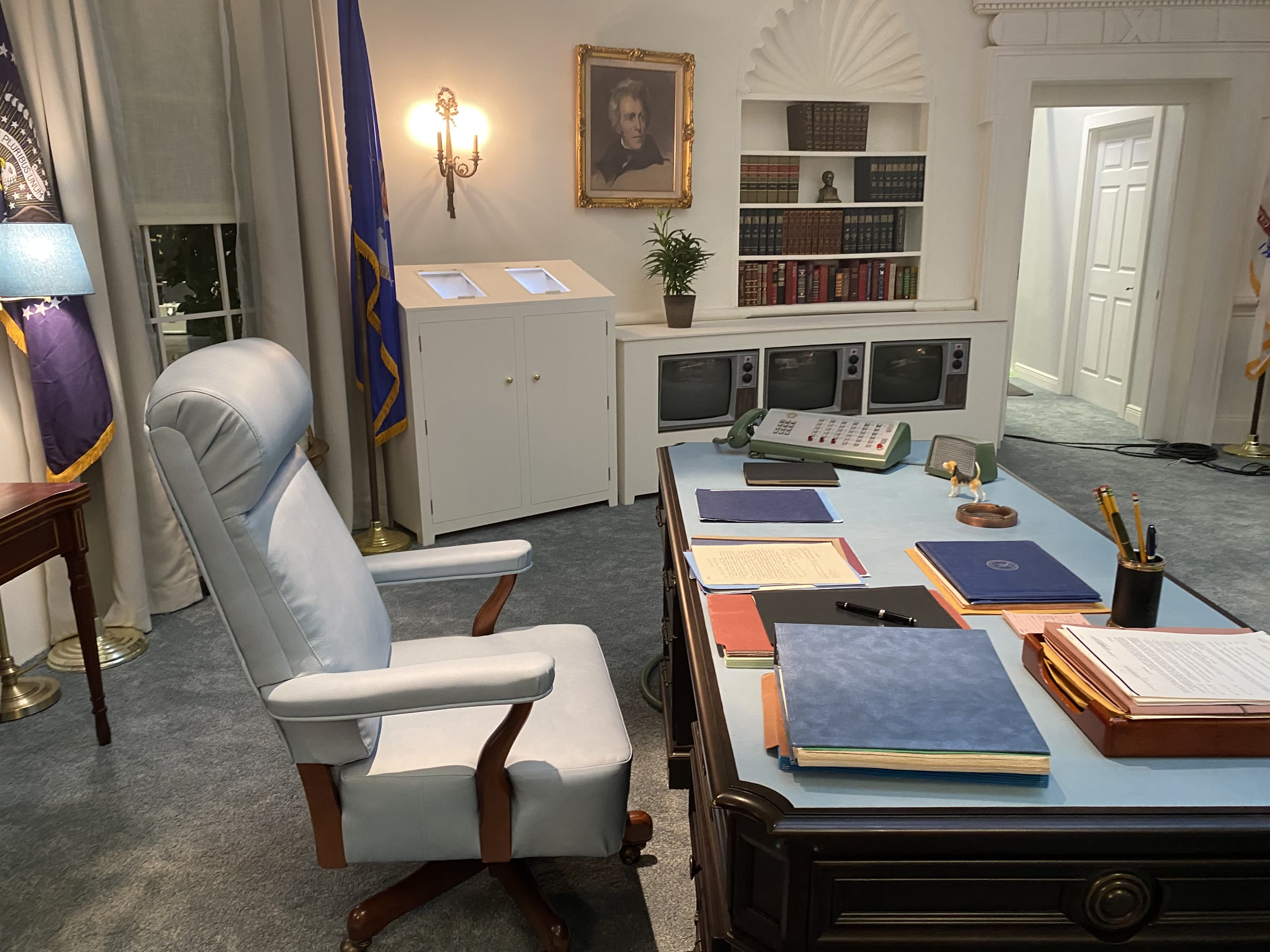 The desk side of the Oval Office set: a three-screen television bank built into a low bookcase, a pale-green multi-button switchboard telephone on the blue leather-topped desk, the president's pale-blue swivel chair, and a white teletype cabinet with two lit angled windows.