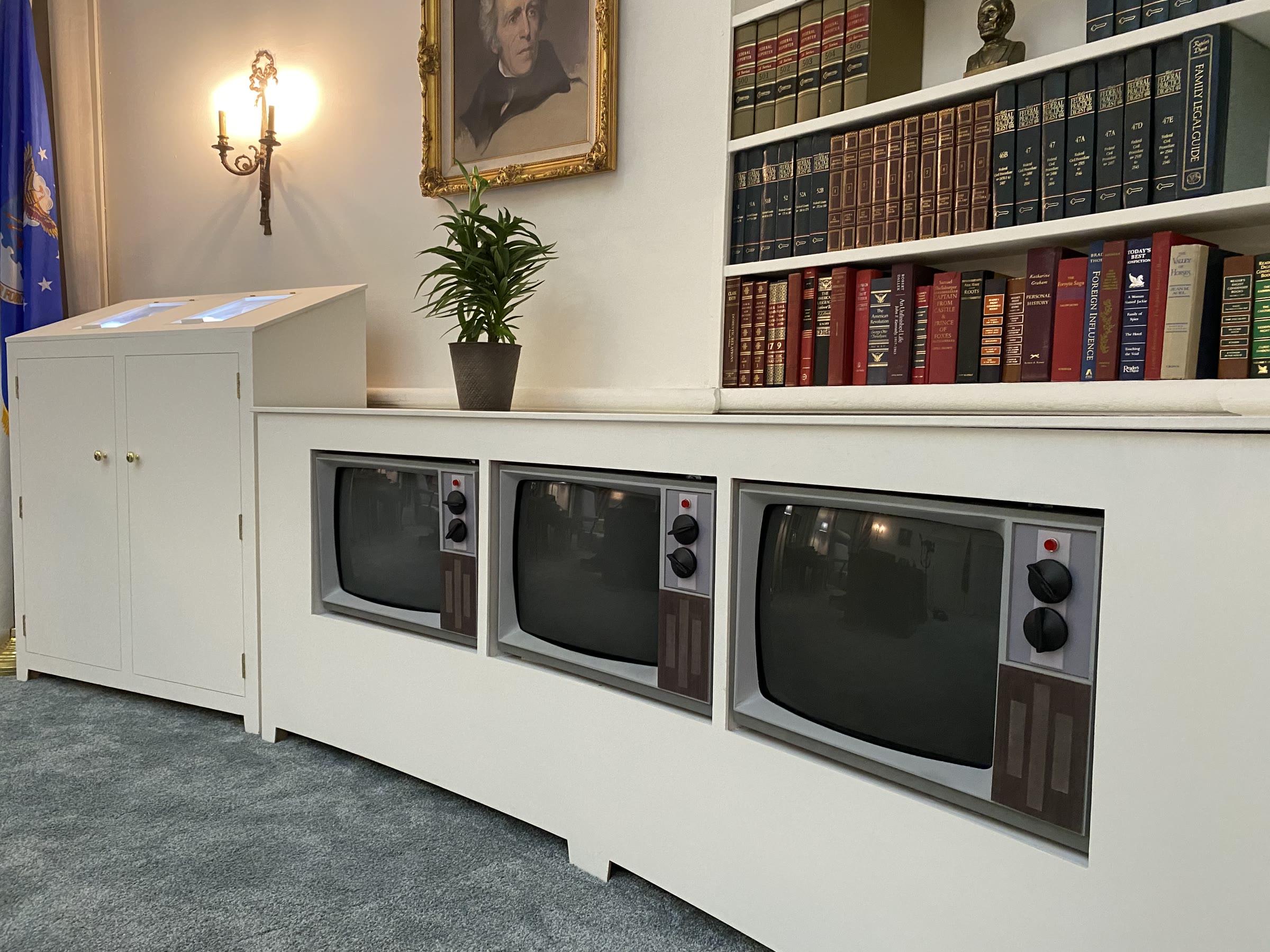 The finished television bank close up: three matched fabricated CRT cabinets recessed in the bookcase with grey bezels, black knobs, and red pilot lights, beside the teletype cabinet's two lit angled windows.