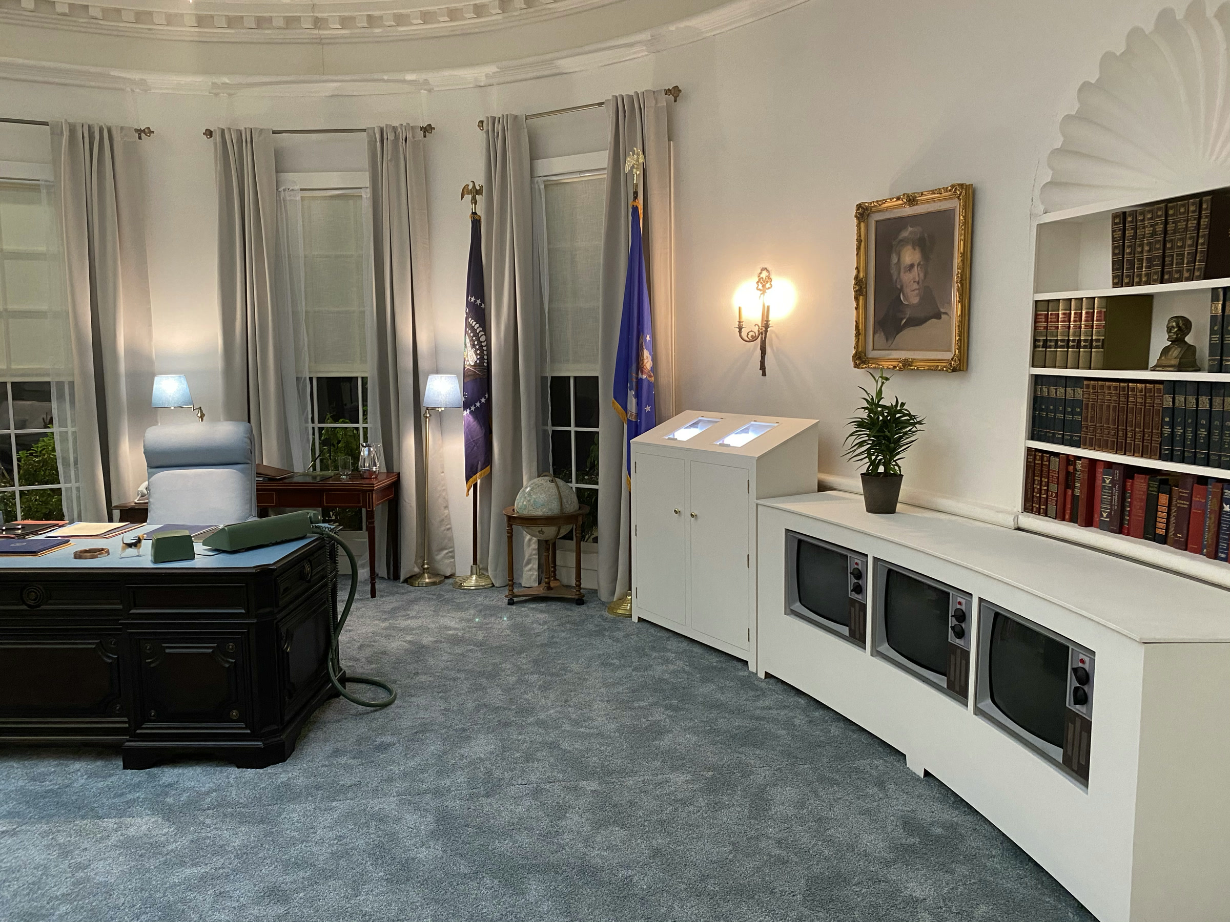 A wide view of the Oval Office set under the oval cove ceiling: curtained windows with flags and a globe, the dark wooden desk with its pale-green switchboard phone, the white teletype cabinet, and the three-screen television bank recessed below the bookcase.