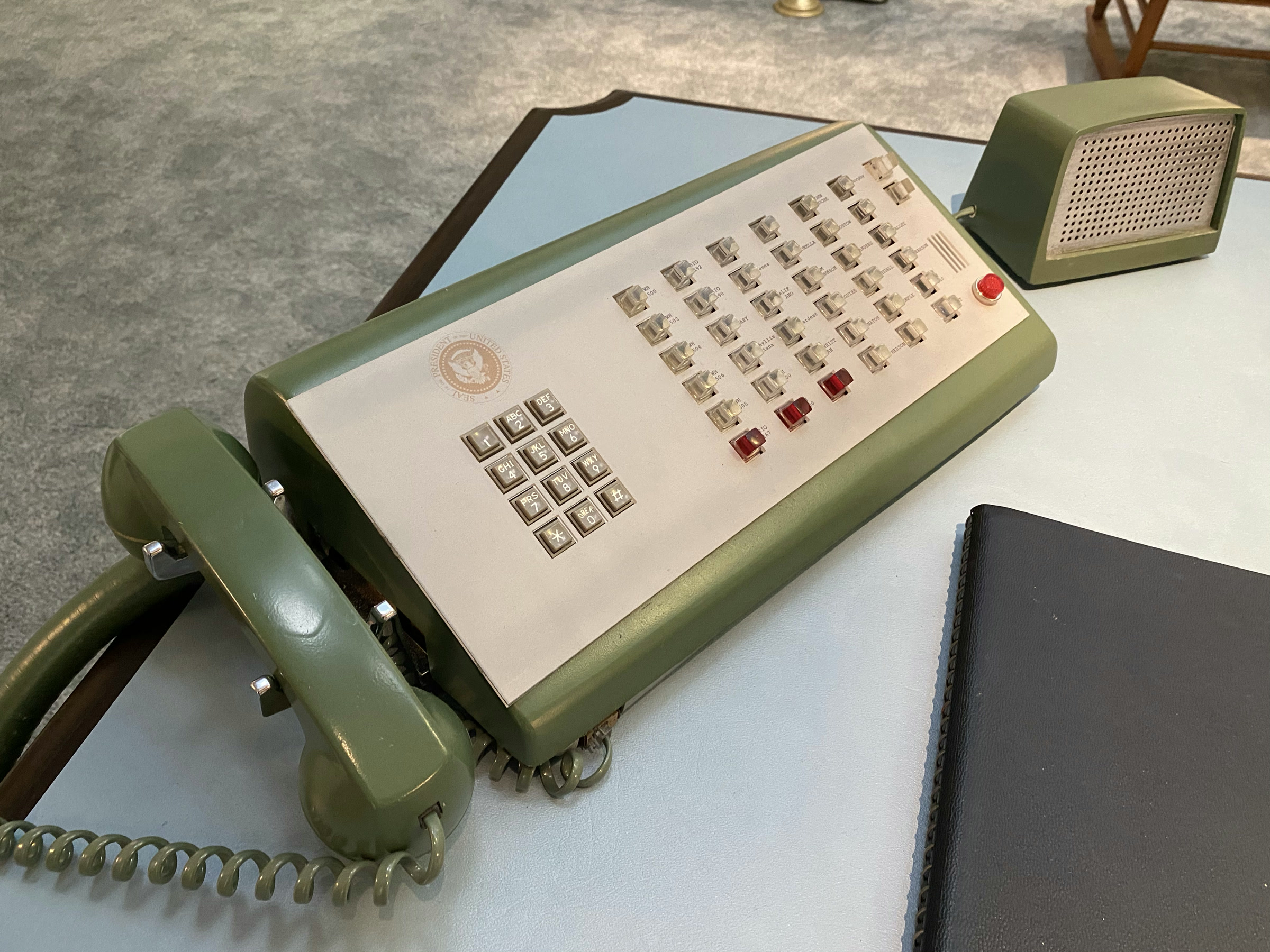 The finished switchboard phone on the blue desk top: pale-green Western Electric body, white face with the presidential seal, rows of lucite buttons with three red, the painted handset, and the matching speaker box behind it.